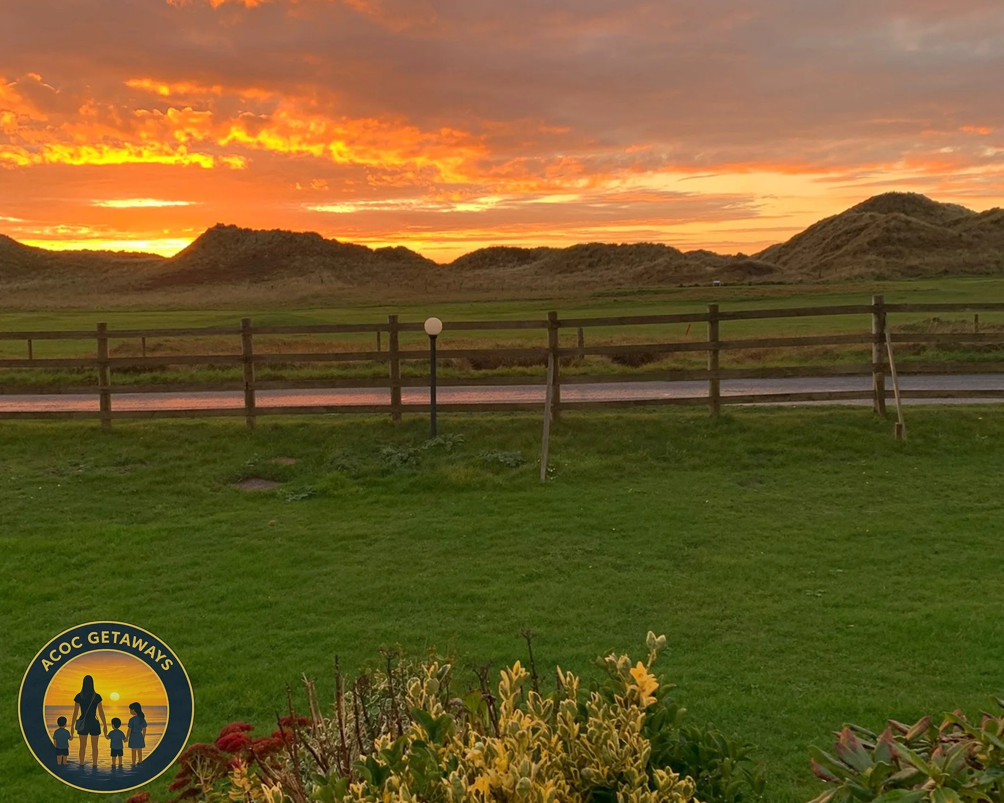 A scenic landscape showing a sunset over grassy hills with a wooden fence, a lamp post, and a patch of blooming flowers in the foreground. The sky is filled with vibrant orange and pink clouds.