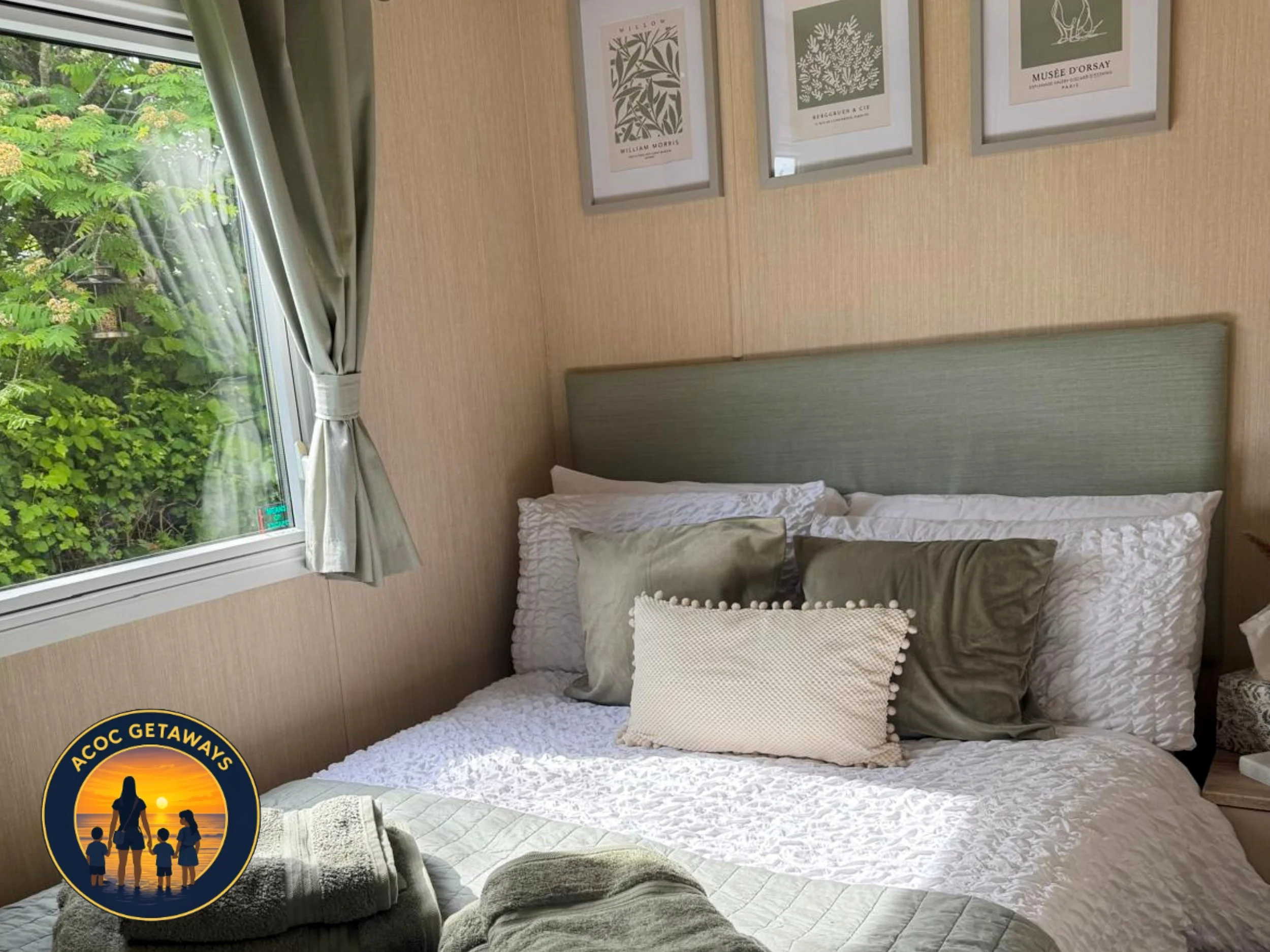 A cozy bedroom with sunlight coming through a window with green trees outside. The bed has white bedding and multiple pillows, including a beige pillow with pom-pom trim. Framed botanical prints hang on the beige wall above the bed. A partial view of