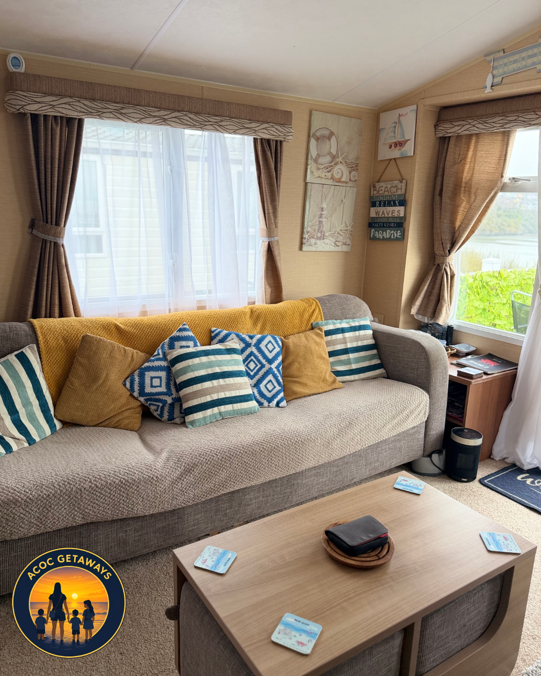Cozy living room with beige sofa adorned with striped and solid pillows, large windows with brown curtains and sheer white curtains, wooden side table with magazines and a black heater, wall decor with beach-themed paintings and a wooden sign, light-