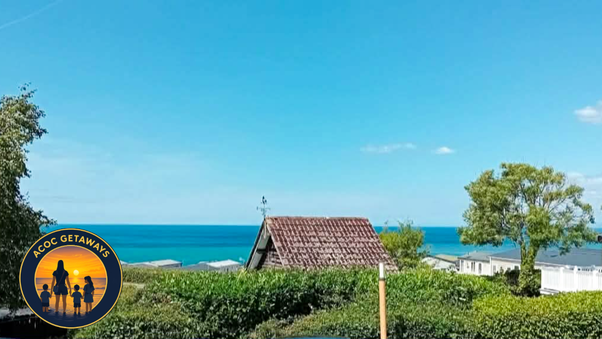 A coastal landscape with a tile-roofed house, green shrubbery, a tall tree, and the ocean in the background under a clear blue sky.