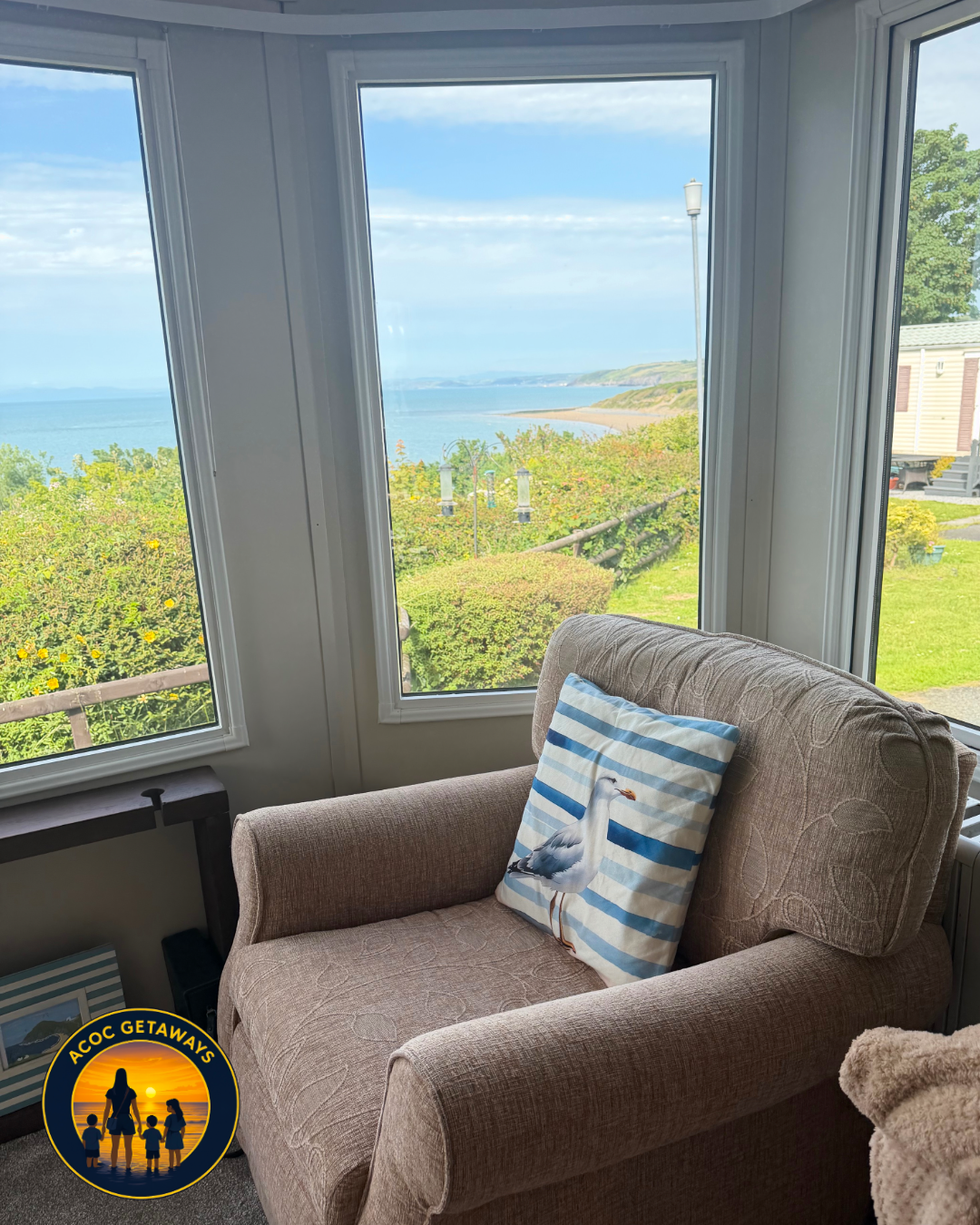 A cozy beige armchair with a decorative pillow featuring a seagull, inside a room with large windows showing a scenic ocean view, green bushes, and a distant coastline under a partly cloudy sky.