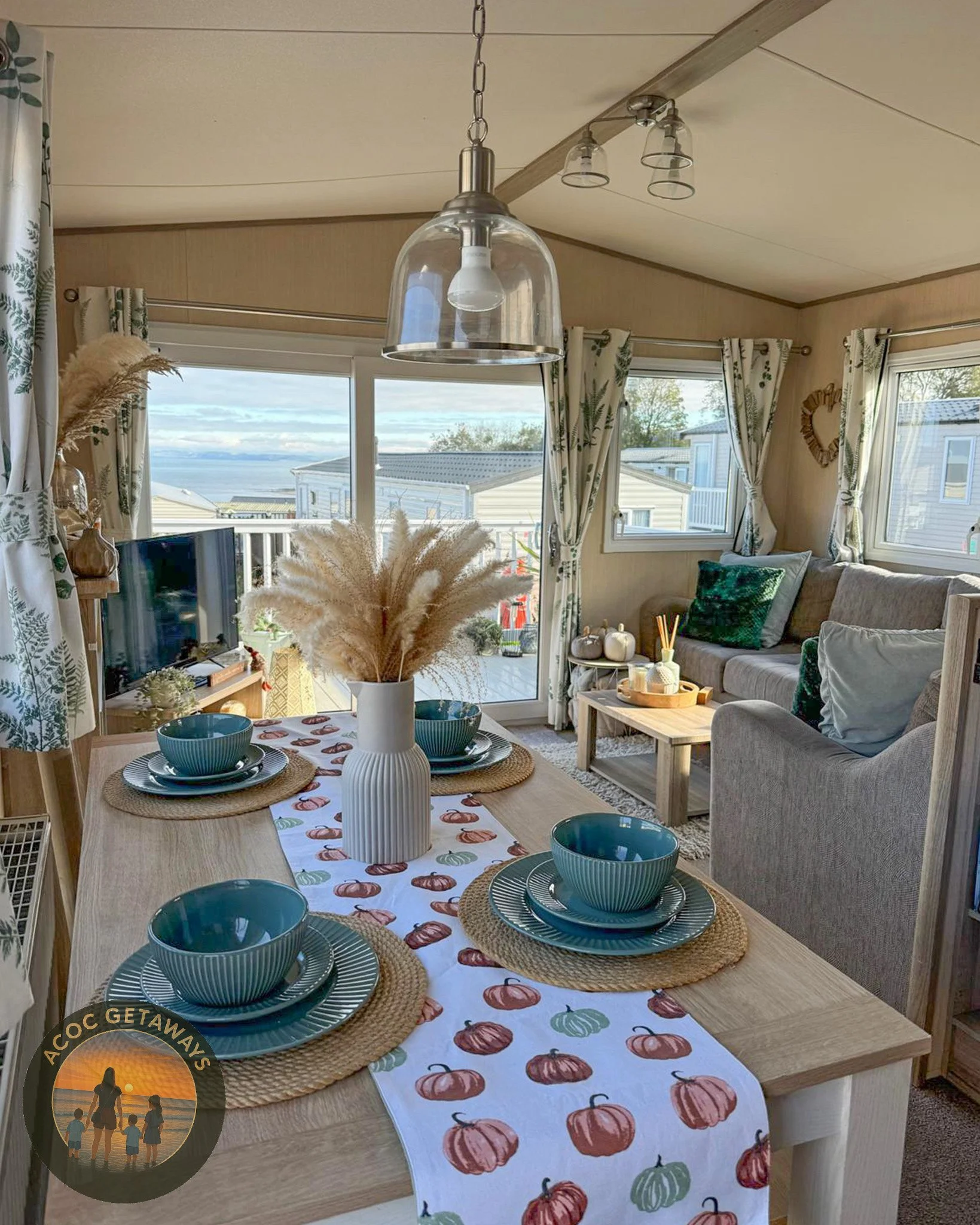 Cozy dining and living area inside a mobile home with large windows showing a view of water and trees. The dining table is set with blue bowls, plates, and woven placemats on a pumpkin-themed table runner. Decor includes pampas grass in a white vase,