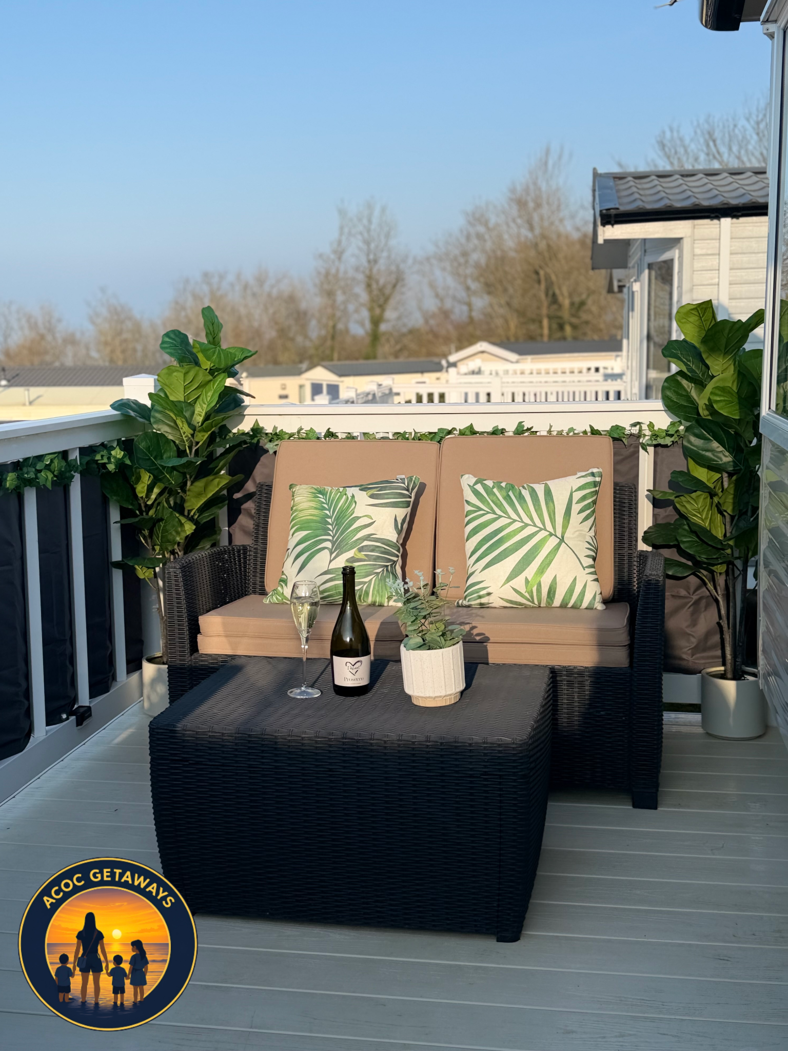 An outdoor balcony with wicker furniture, pillows with tropical leaf patterns, a bottle of wine, a glass, and potted plants, with a view of neighboring rooftops and trees under a clear blue sky.