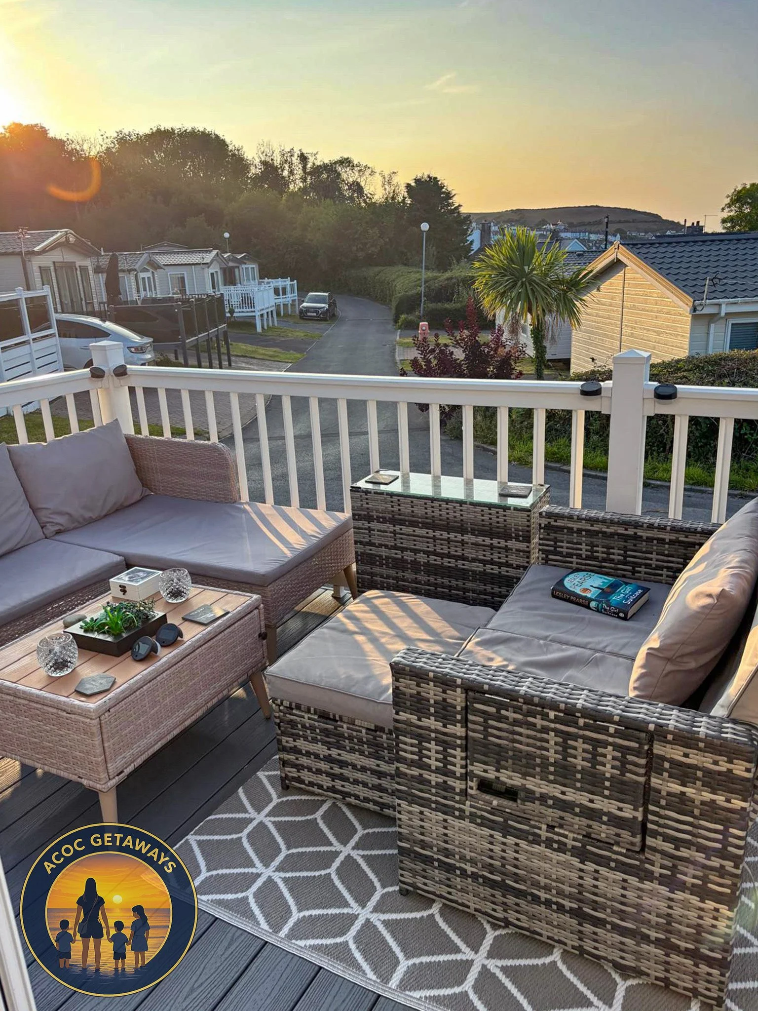 A balcony with outdoor wicker furniture including chairs, a sofa, and a glass-top table with decorative items and a book, overlooking a residential neighborhood during sunset.
