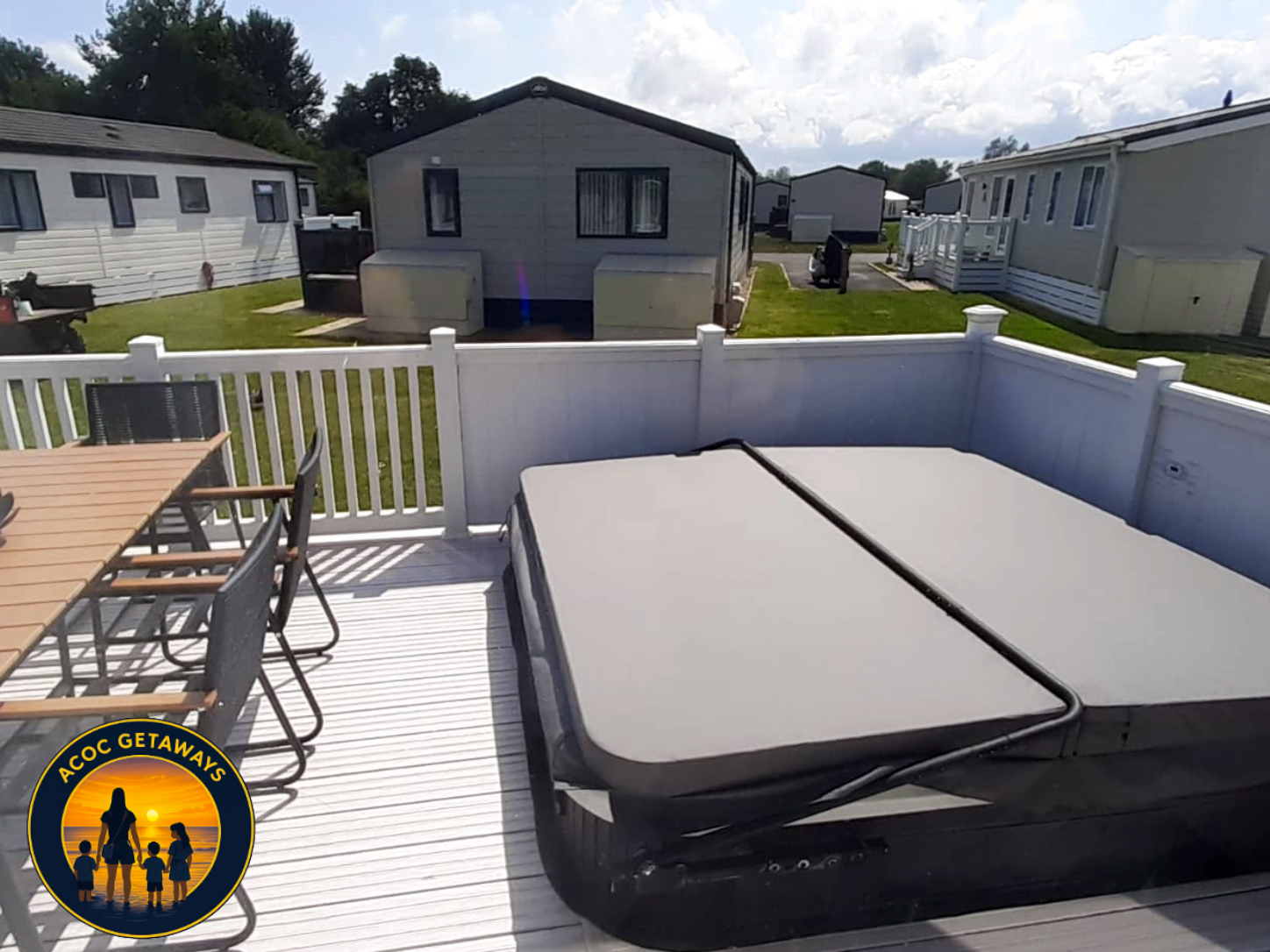 View from a screened porch showing outdoor furniture, a hot tub, and a row of mobile homes or cabins in a park-like setting with green grass, trees, and a partly cloudy sky.