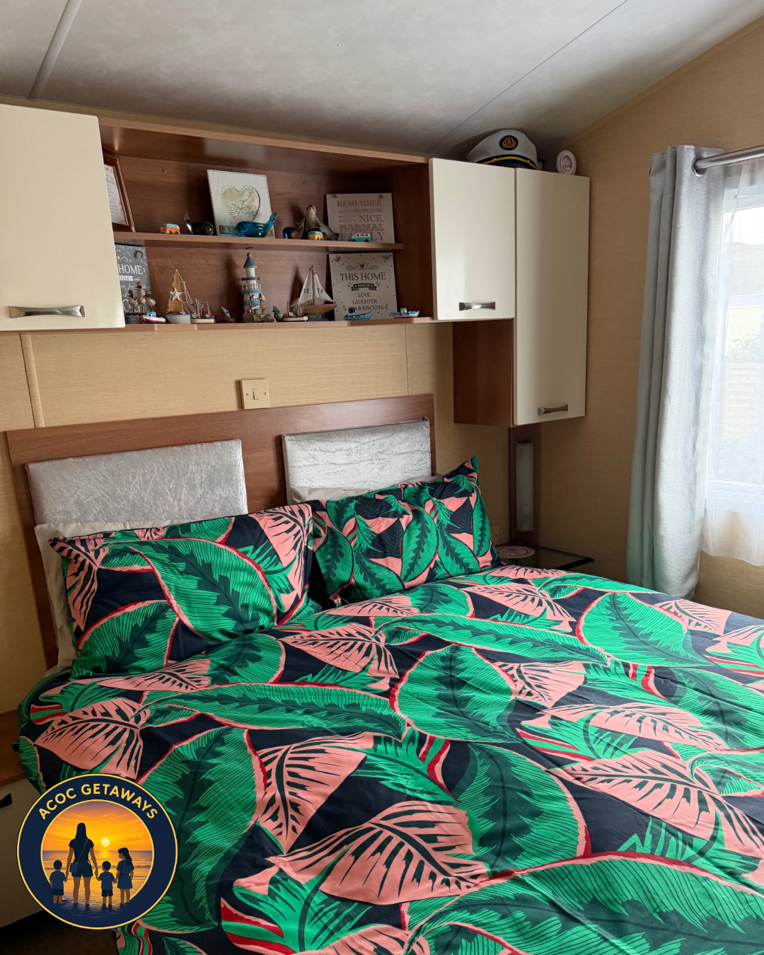 A bedroom with a bed featuring a colorful tropical leaf patterned comforter and pillows. There are built-in wall cabinets and shelves with decorative items, including nautical-themed décor. A cap rests on top of the cabinet. Natural light comes throu