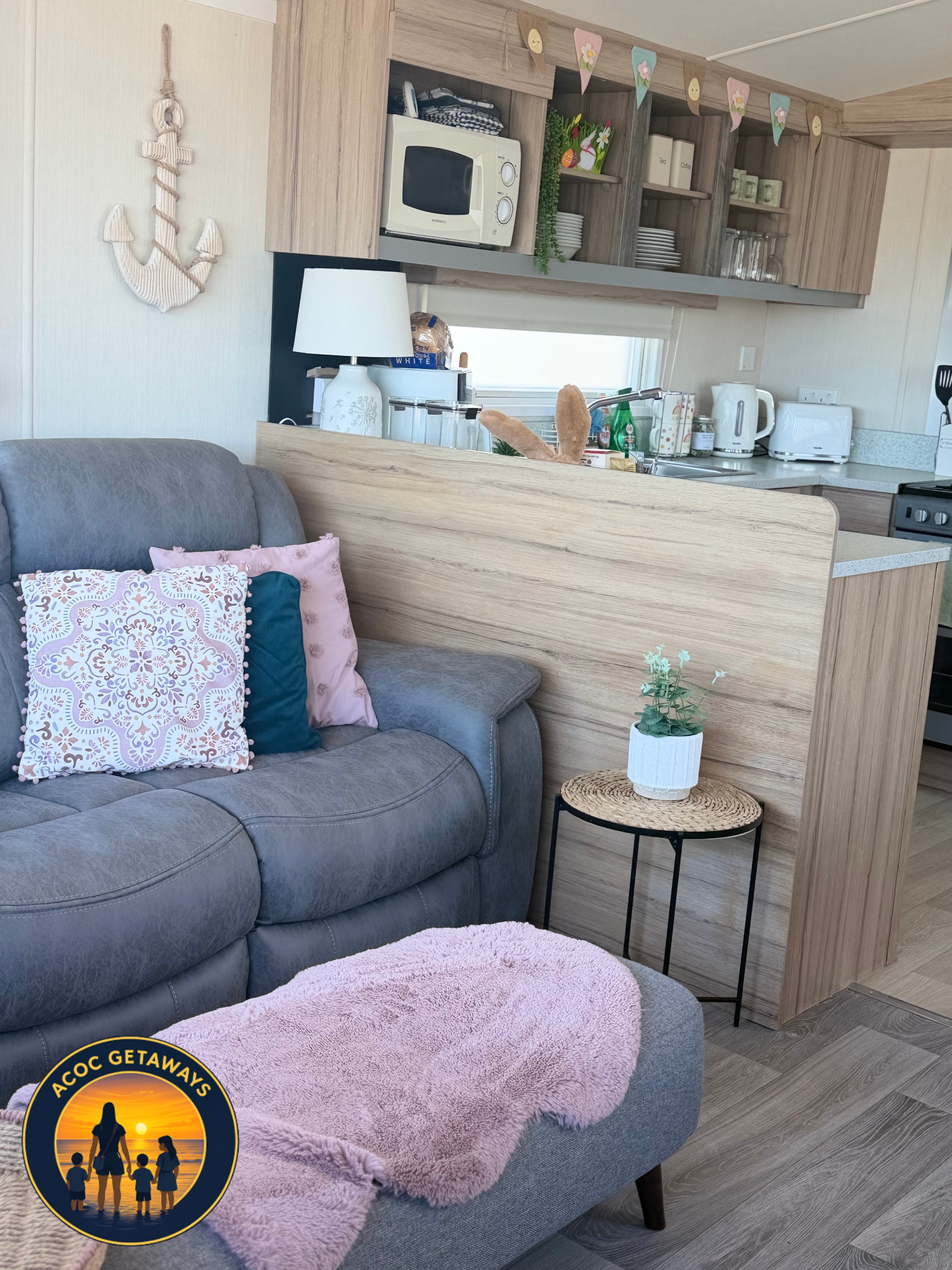 Living room with gray couch, pink throw blanket, decorative pillows, small side table with a potted plant, visible kitchen area with microwave, kettle, toaster, and bunting decorations hanging above.