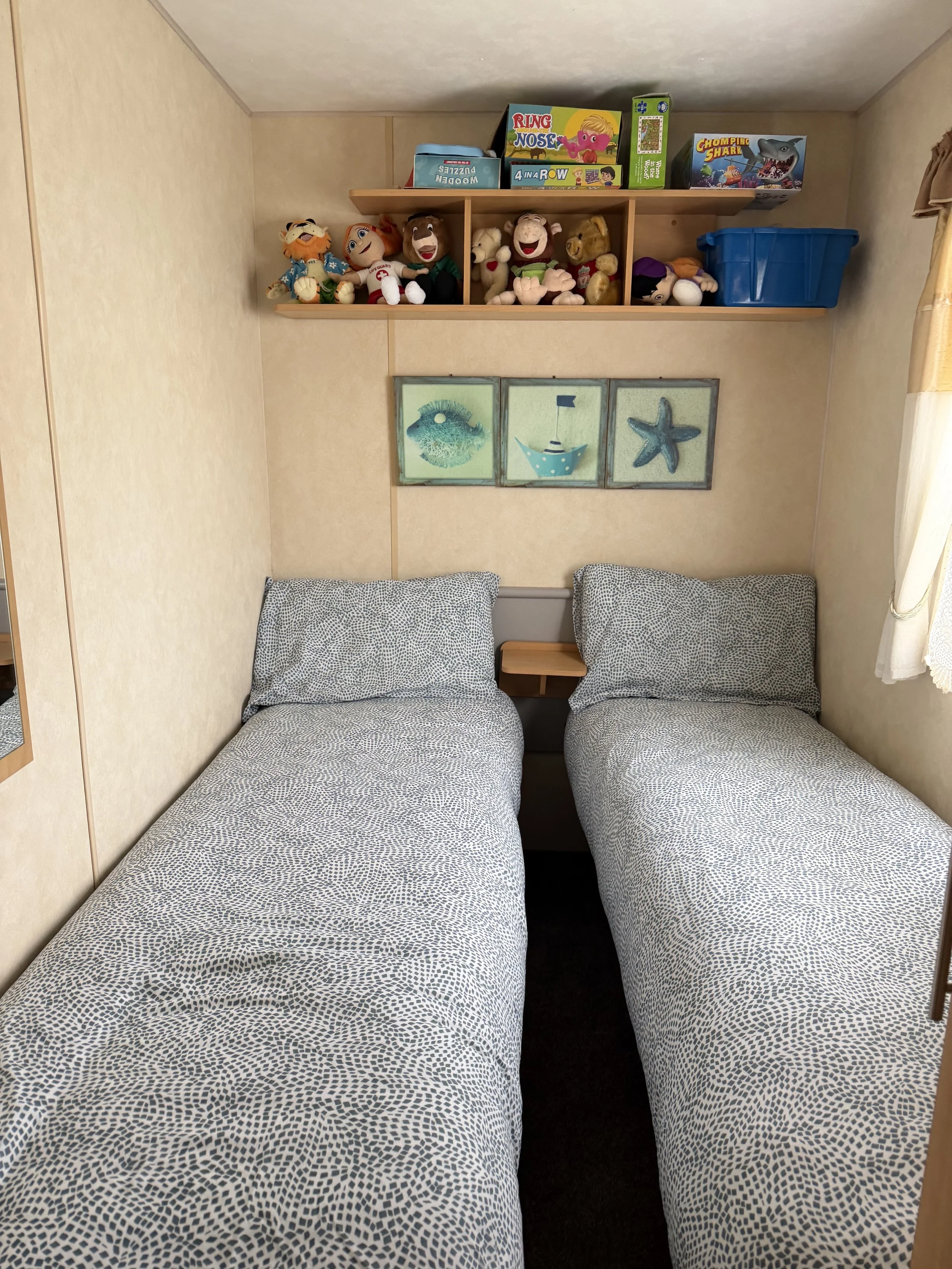 Two twin beds with matching patterned bedspreads in a small bedroom, with a small nightstand between them, two pillows, a wall shelf holding plush toys and board games above the beds, and framed nautical-themed pictures on the wall.