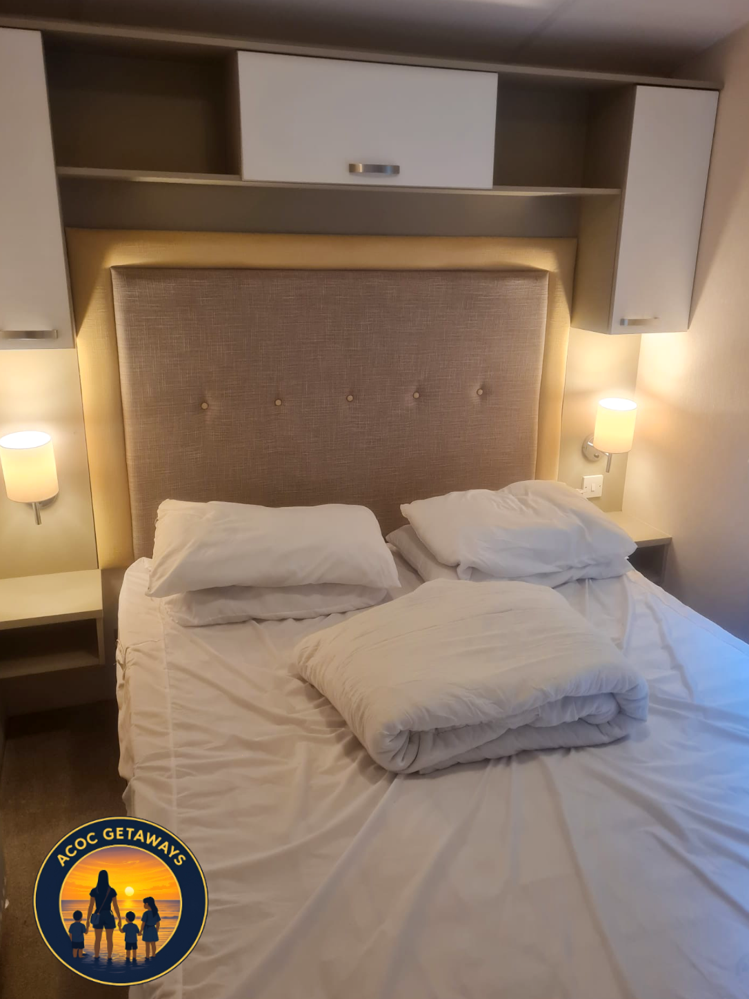 Unmade bed with pillows and blankets, beige padded headboard, wall-mounted lamps on either side, and built-in shelves and cabinets above the bed in a hotel room.
