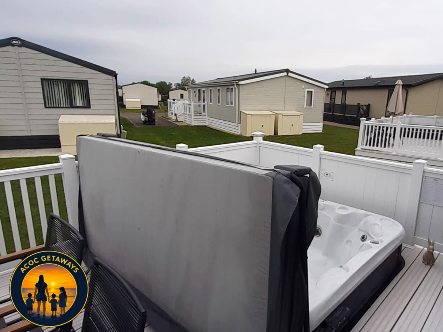 View of above-ground hot tub on a deck with a privacy screen, surrounded by a white fence, with neighboring houses and yards visible under cloudy sky in a residential park.
