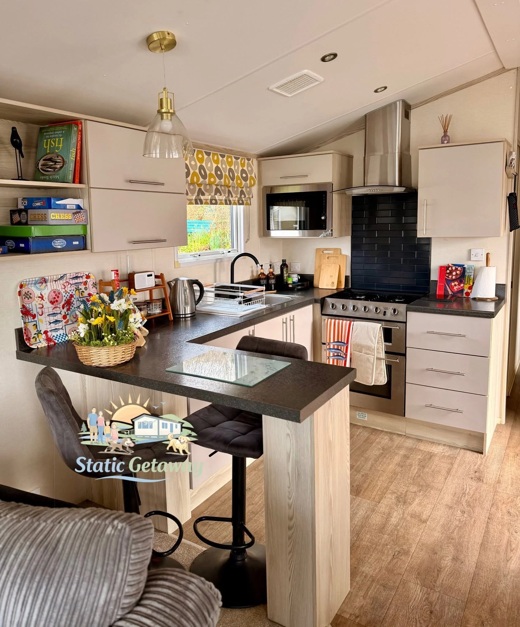 A modern kitchen with light wood cabinets, a black countertop, and stainless steel appliances including a microwave and oven. There is a small window with a patterned valance, black range hood, and various kitchen items on the counter. A small family