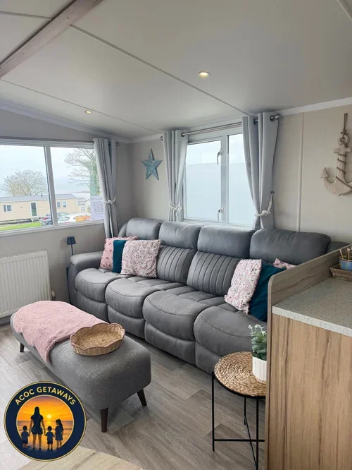 Living room with gray sectional sofa, pink and teal pillows, soft pink blanket on ottoman, wicker basket, small side table, and coffee plant, with large windows, white curtains, and coastal decor in the background.