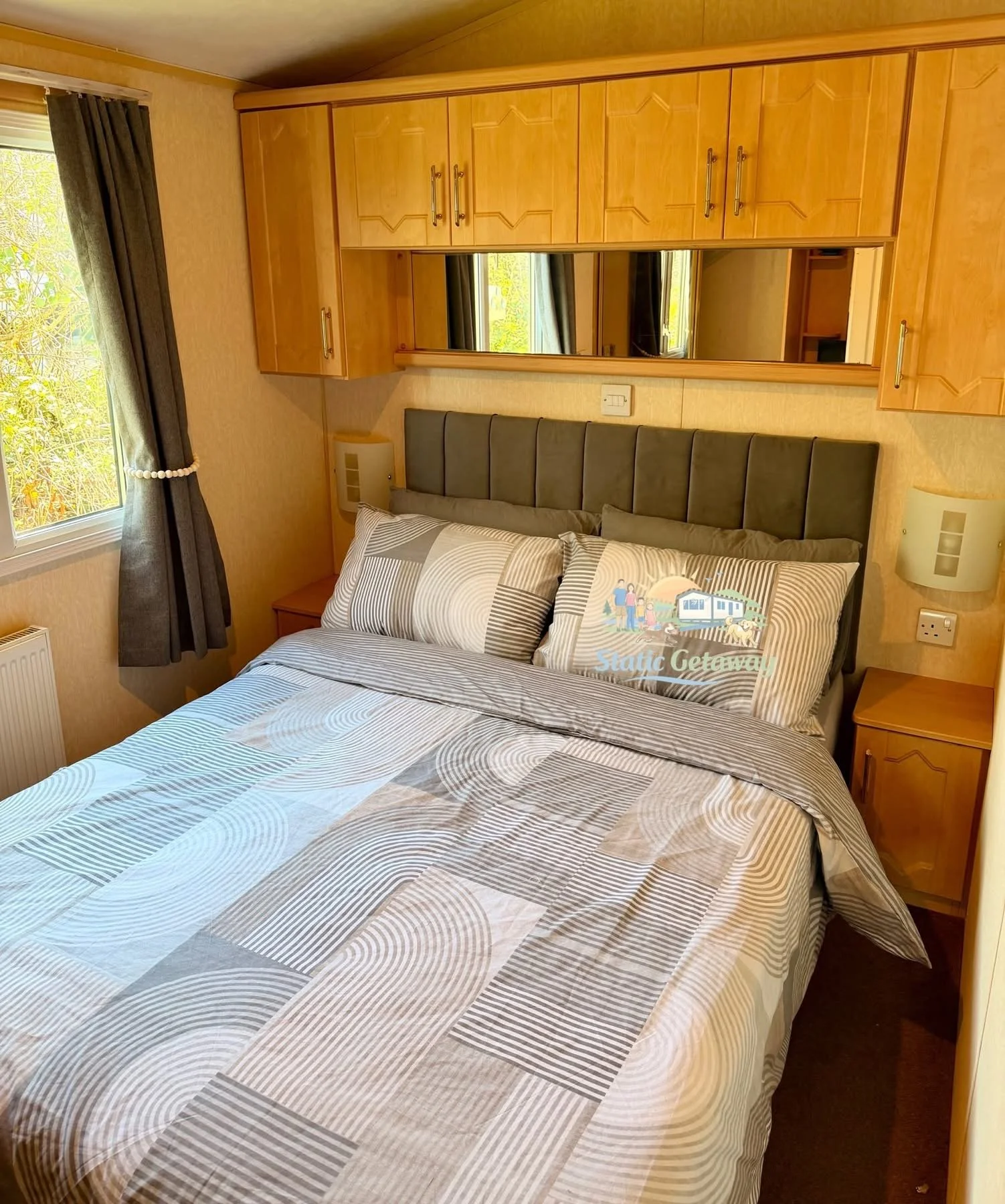A cozy bedroom with a bed covered in geometric patterned bedding. The bed has a padded headboard and two matching pillows. Behind the bed is a large mirror with wooden cabinets above it. To the left, a window with dark curtains and a view of greenery