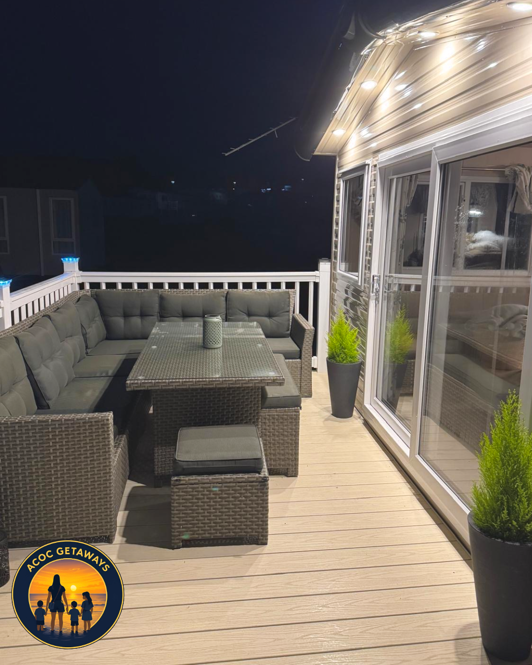 Nighttime outdoor patio with wicker sectional sofa, glass-top table, potted green plants, and sliding glass doors leading inside.