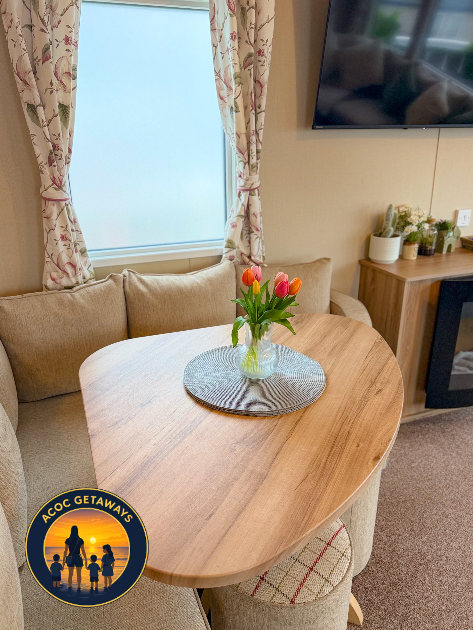 A cozy living room corner with a wooden dining table, a beige cushioned chair, a floral curtain, a TV mounted on the wall, and a vase of pink and orange tulips on the table.