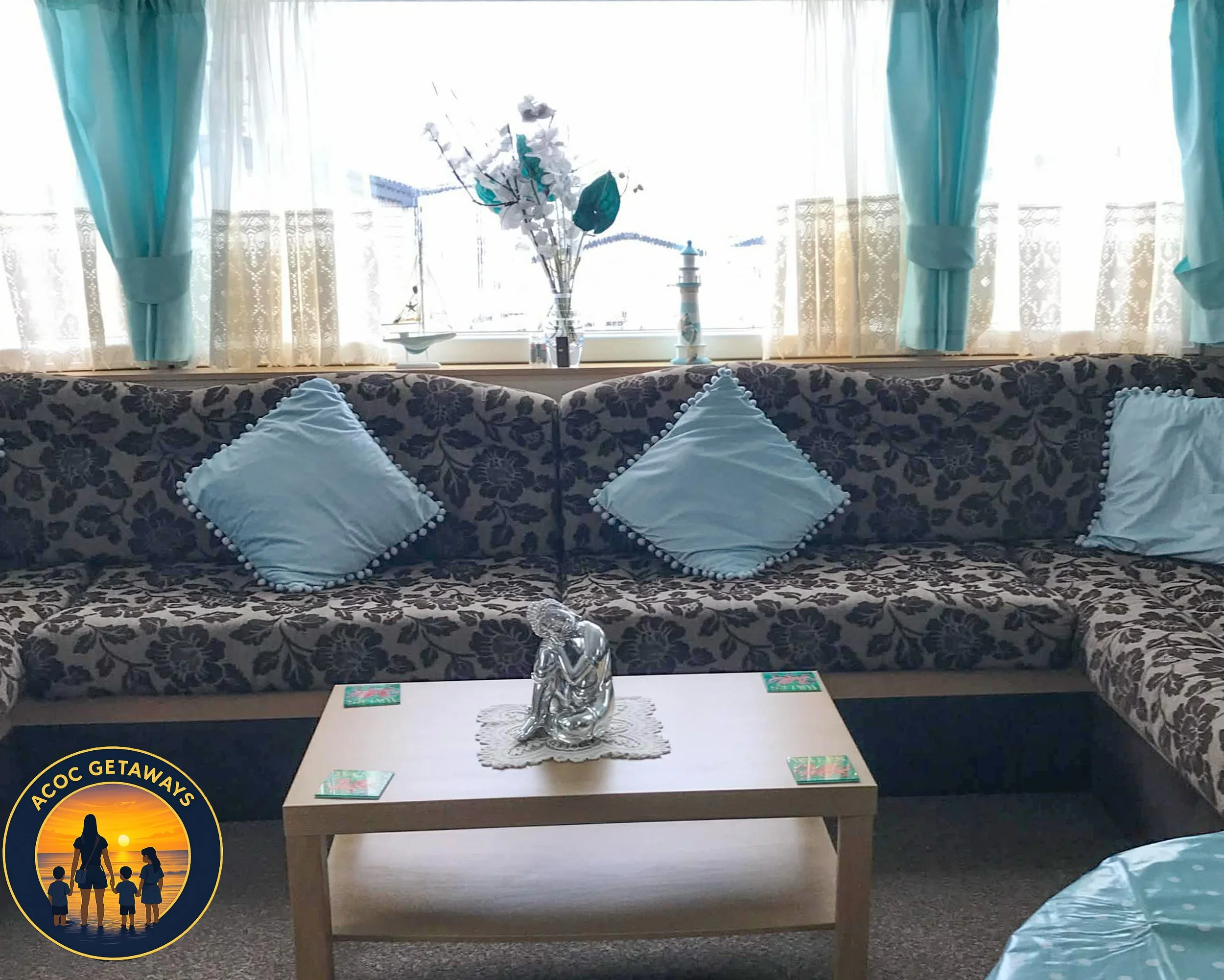 Living room with a patterned sectional sofa decorated with light blue pillows, a coffee table with a decorative sculpture, window with sheer curtains and blue drapes, and decorative items on the window ledge.