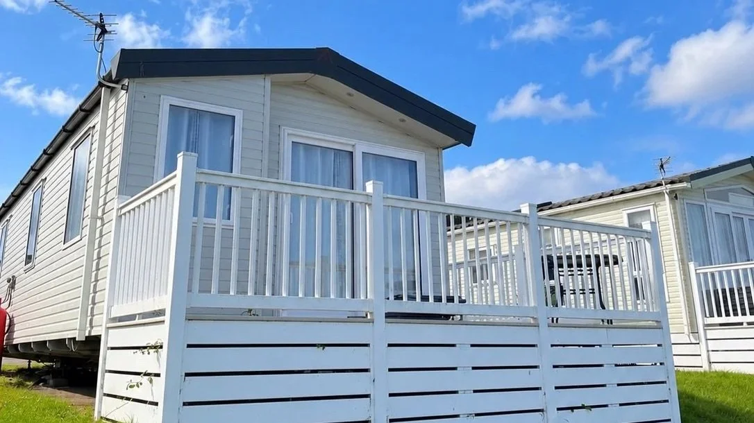 A white house with a small balcony railing, large windows, and a green grassy yard underneath a partly cloudy blue sky.