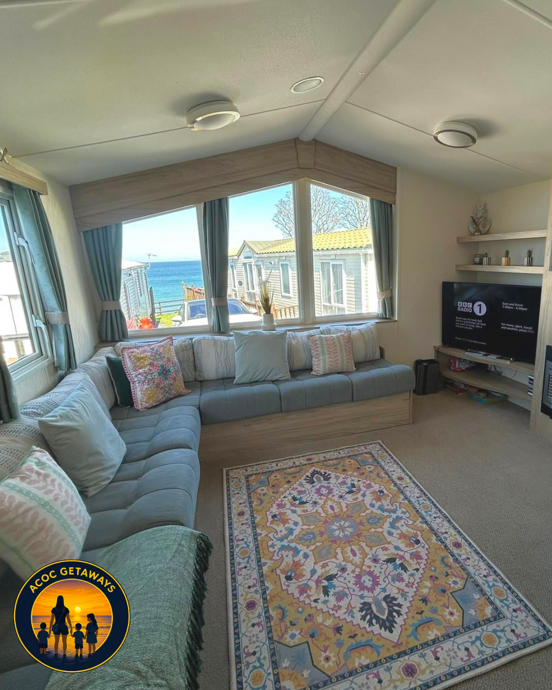 Living room with large windows showing ocean view, light blue sectional sofa with pillows, patterned area rug, TV on a wall shelf, and beige carpet.