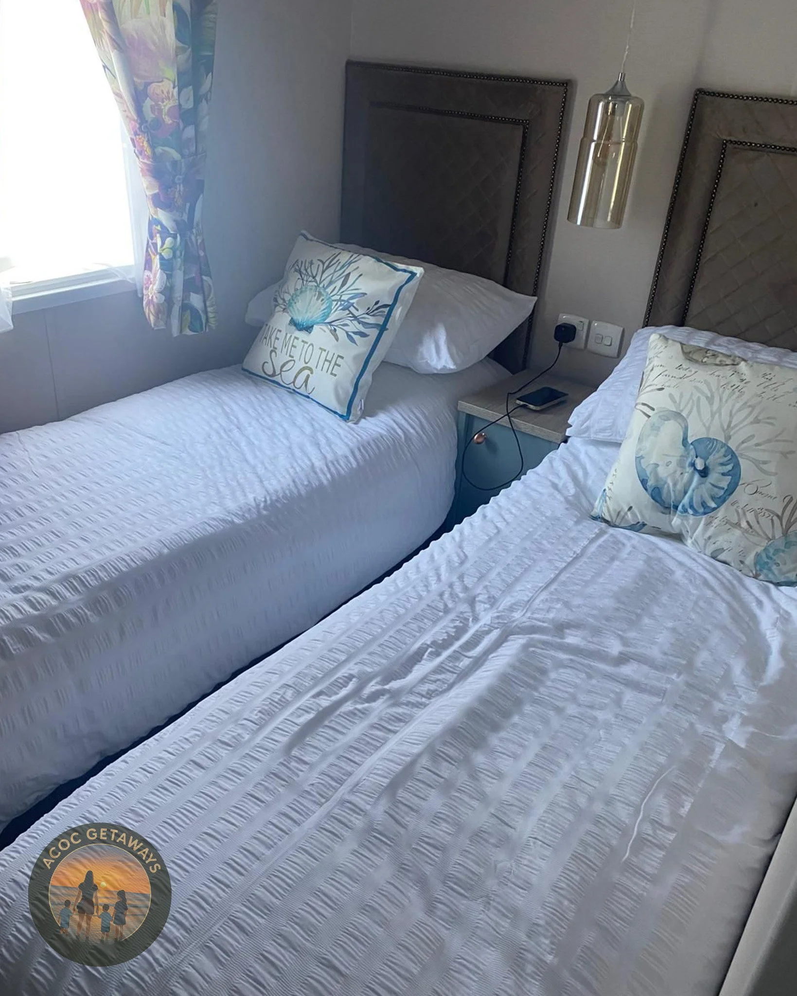 Two single beds with white bedspreads and pillows, one with a seashell themed pillow, are next to a small nightstand with a phone and charging cable. A window with floral curtains is on the left, and there are two headboards with dark upholstery and 