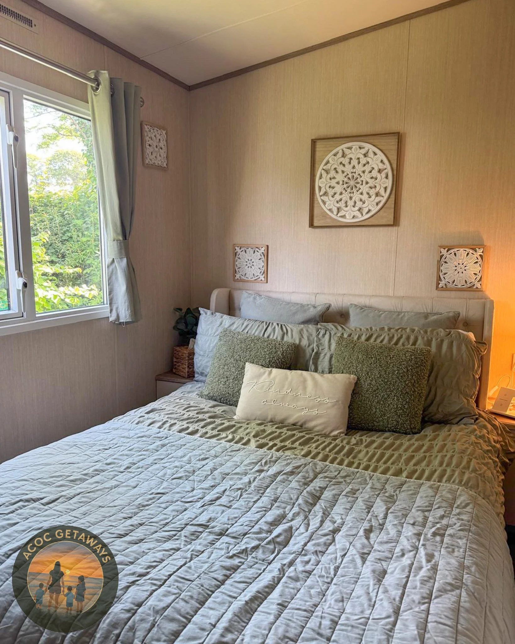 A cozy bedroom with a bed featuring multiple pillows, a window with green foliage outside, decorative wall art above the bed, a nightstand with a basket and a plant, and a warm lamp light.