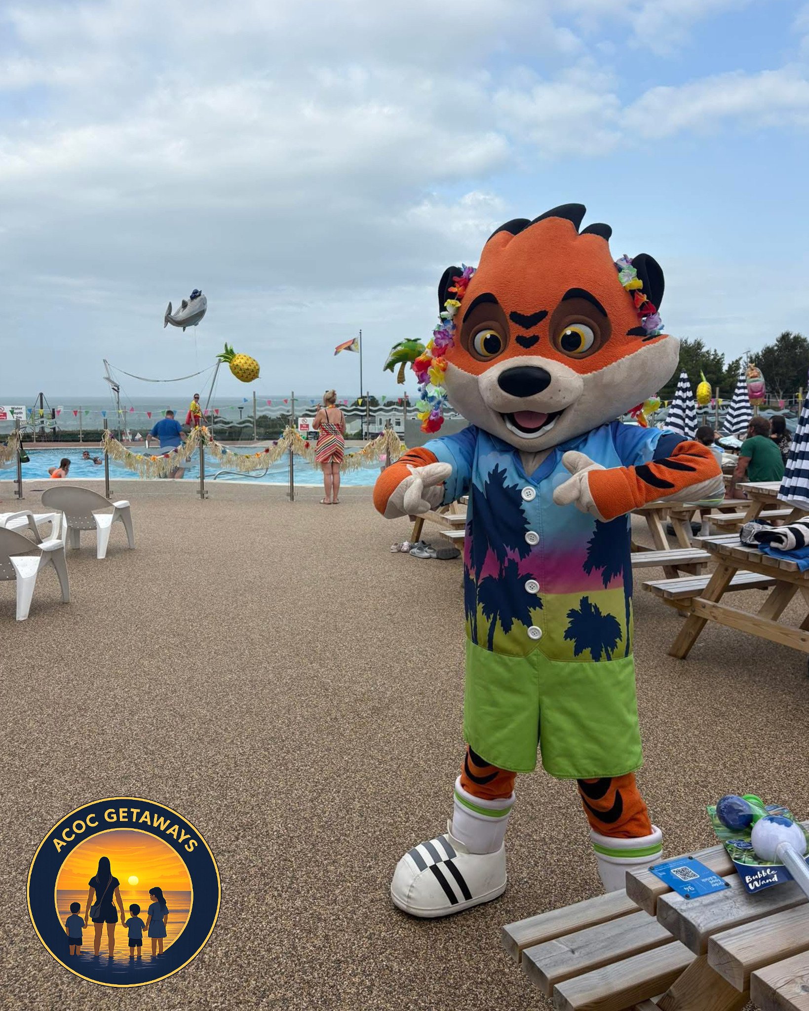 Costumed character of an orange tiger wearing a colorful Hawaii shirt and green shorts at a poolside area with people and a pool in the background.