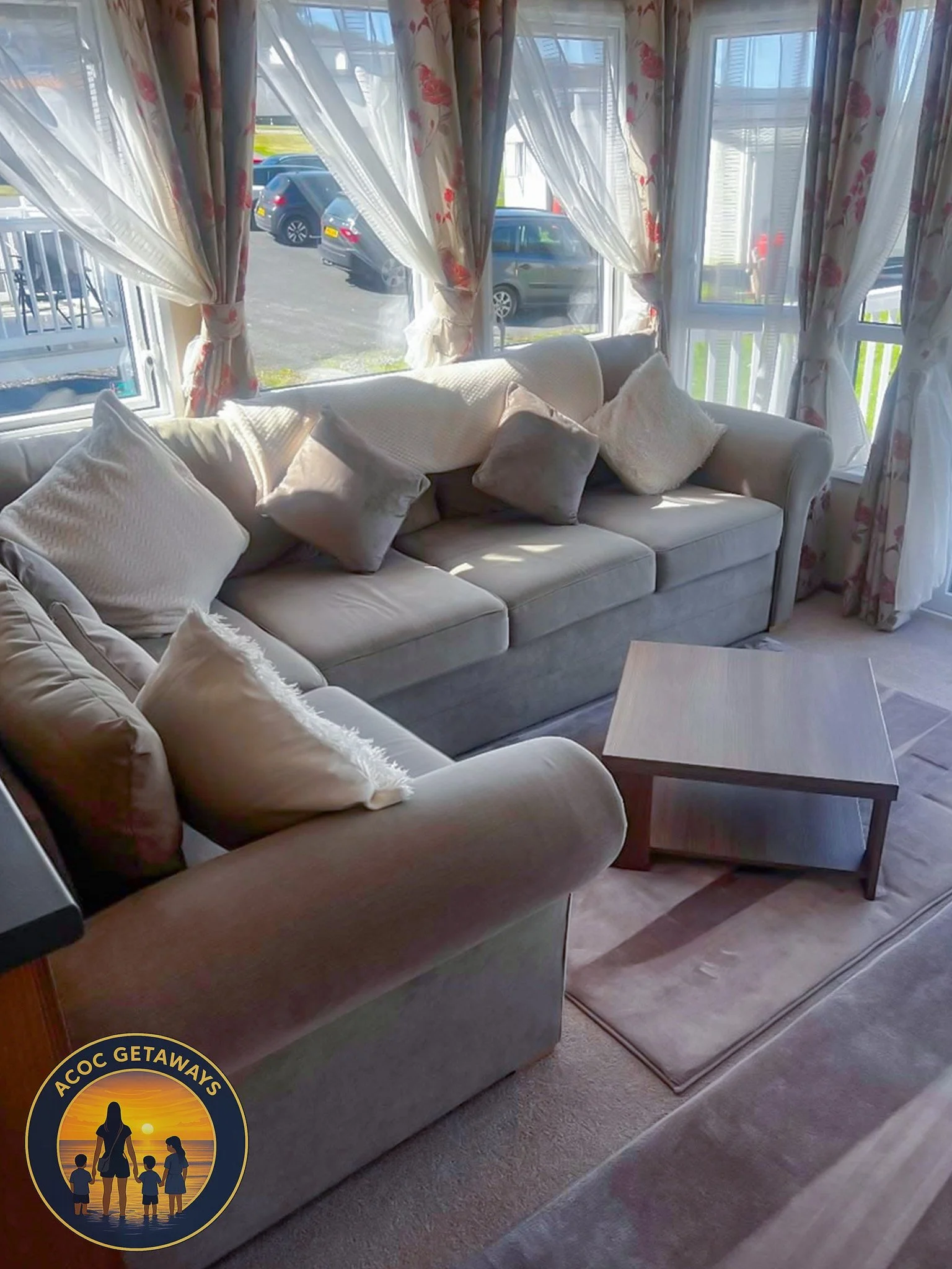 Sunroom with large windows, floral curtains, a white sofa with multiple pillows, a beige armchair with a pillow, a wooden coffee table, and sunlight streaming through the windows.