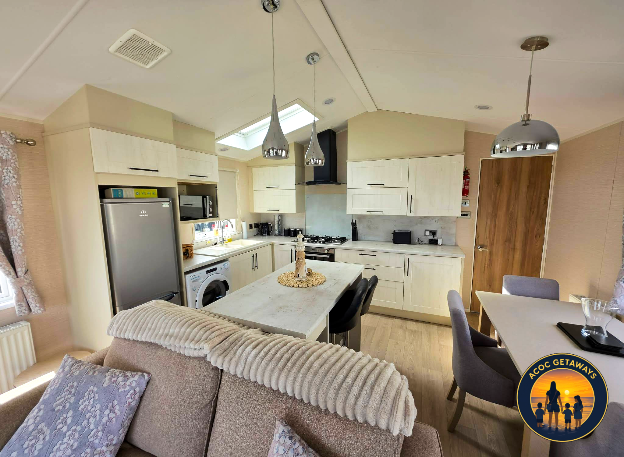 A kitchen with white cabinets, a gray refrigerator, and hanging pendant lights. There is a skylight in the ceiling providing natural light, and a washing machine is visible near the sink. A dining table with black chairs is in the foreground, and par