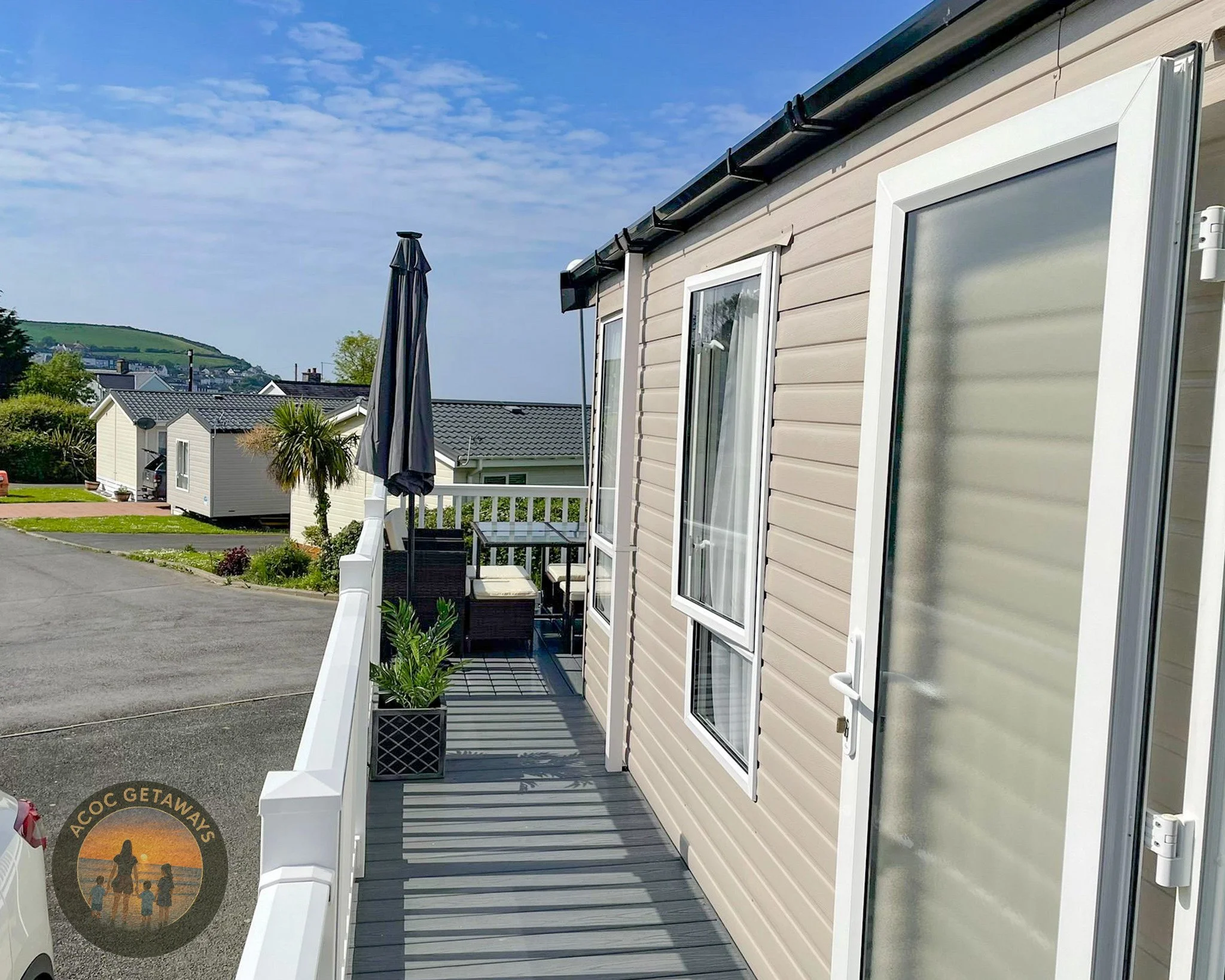 View of a mobile home porch with outdoor furniture, umbrella, and potted plants, overlooking a residential area with other mobile homes and green hills in the background.