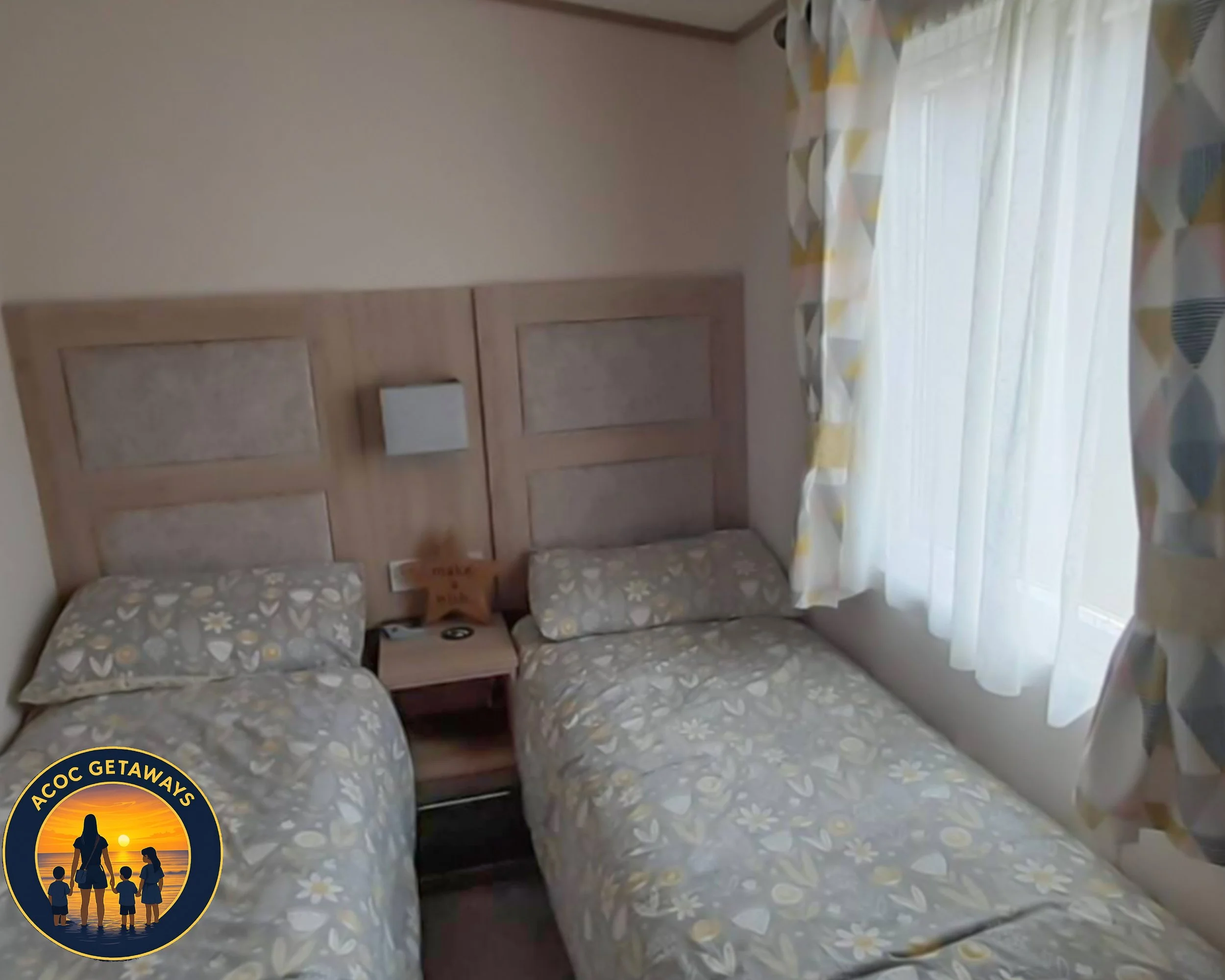 Hotel room with two single beds, a nightstand between them, and a window with patterned curtains. The beds have floral patterned bedding and there is a star-shaped pillow on the nightstand.