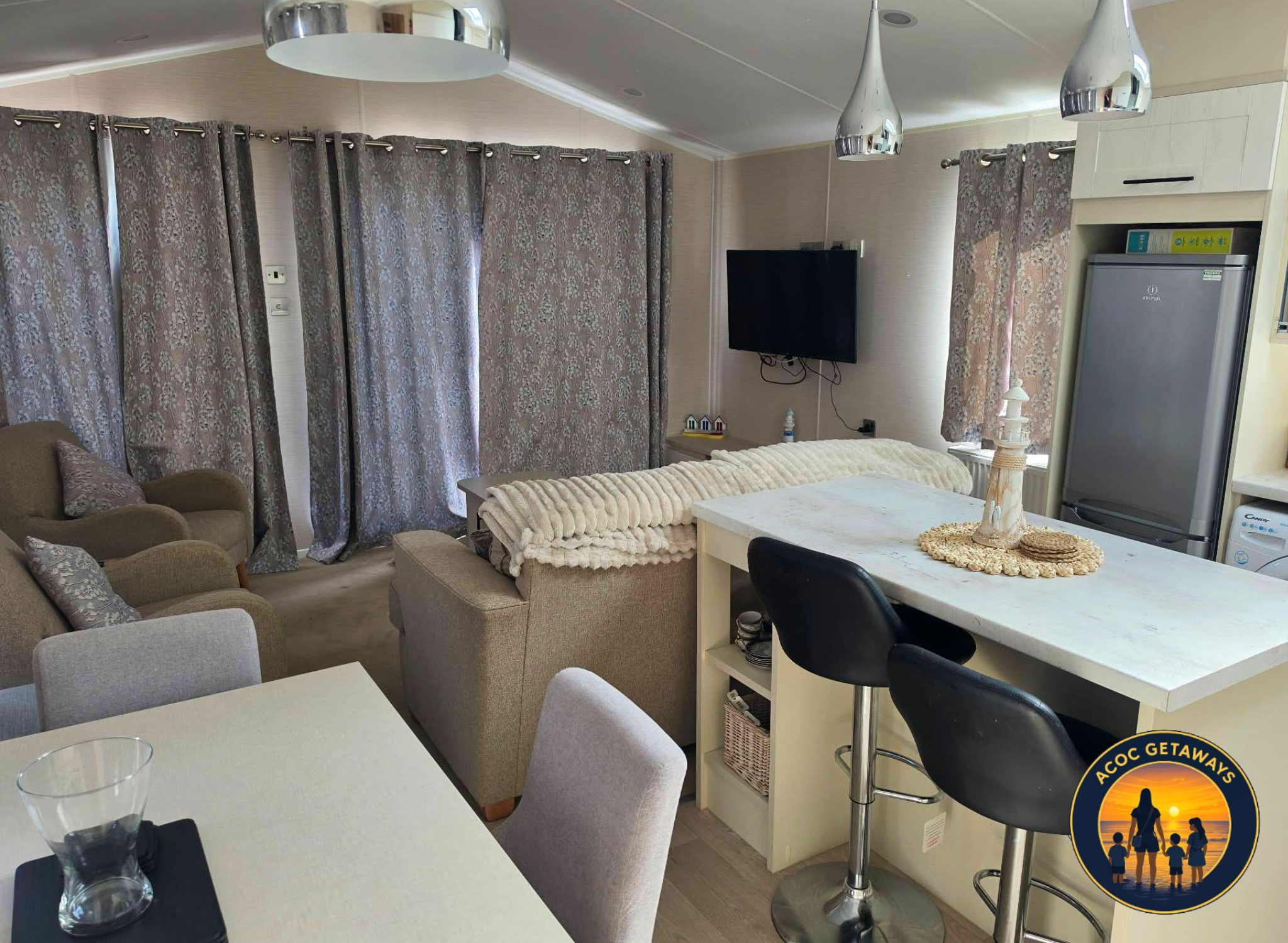 Interior of a living room with beige sofas, patterned curtains, a wall-mounted flat-screen TV, and a dining table with glasses and chairs. There is a kitchen area with a white countertop, black bar stools, and a refrigerator. Decor includes a lamp, s