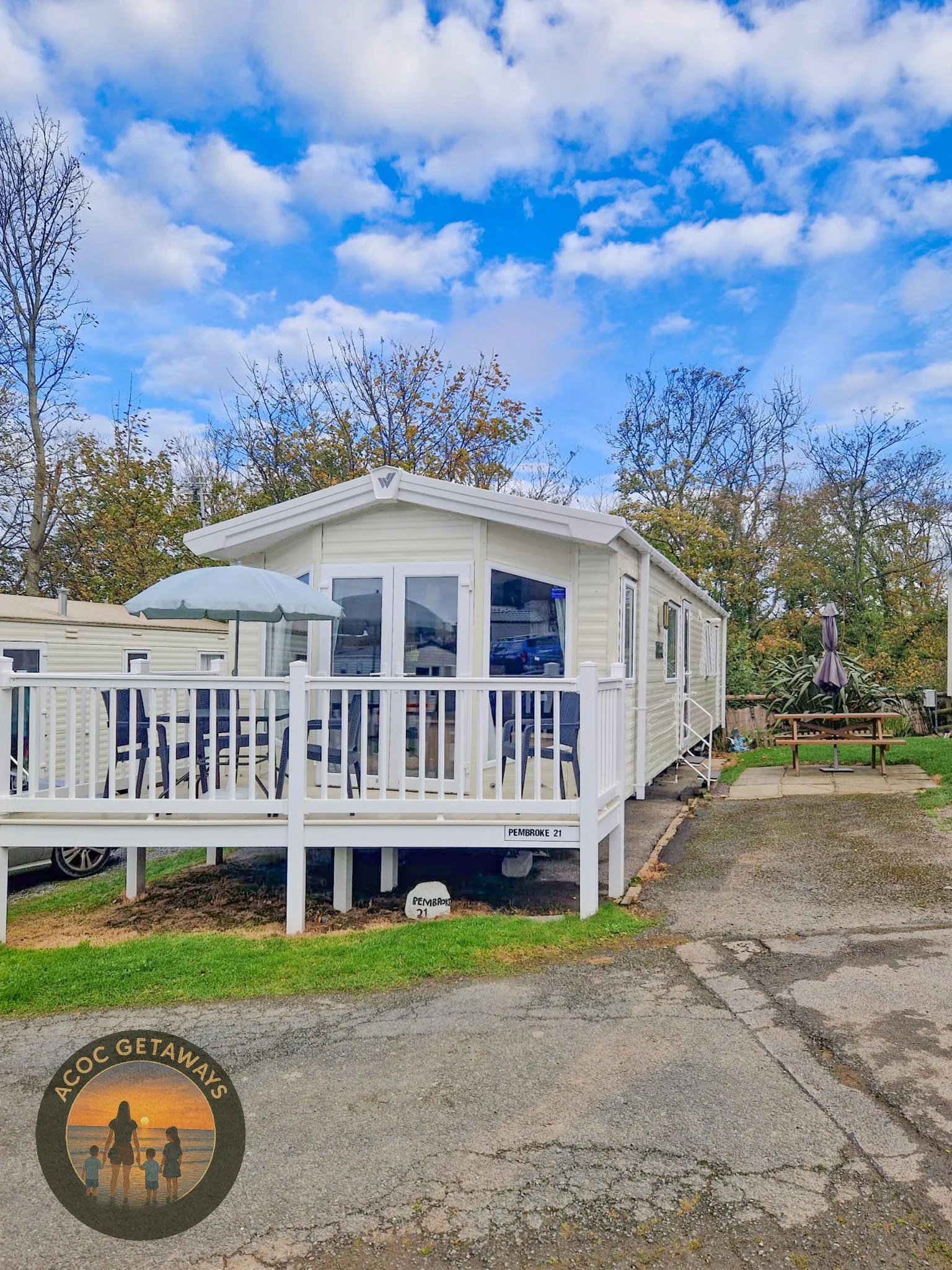 A white mobile home or trailer with a porch, outdoor table with chairs, and patio umbrella, located on a paved pathway with grass and trees in the background under a blue sky with clouds.