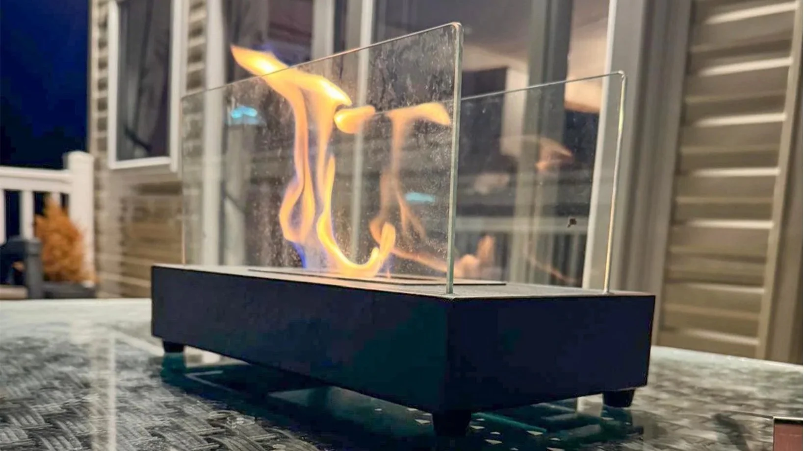 A tabletop fire pit with flames behind a clear glass barrier, set on a dark rectangular base on an outdoor table at night, with blurred background of a house and porch.