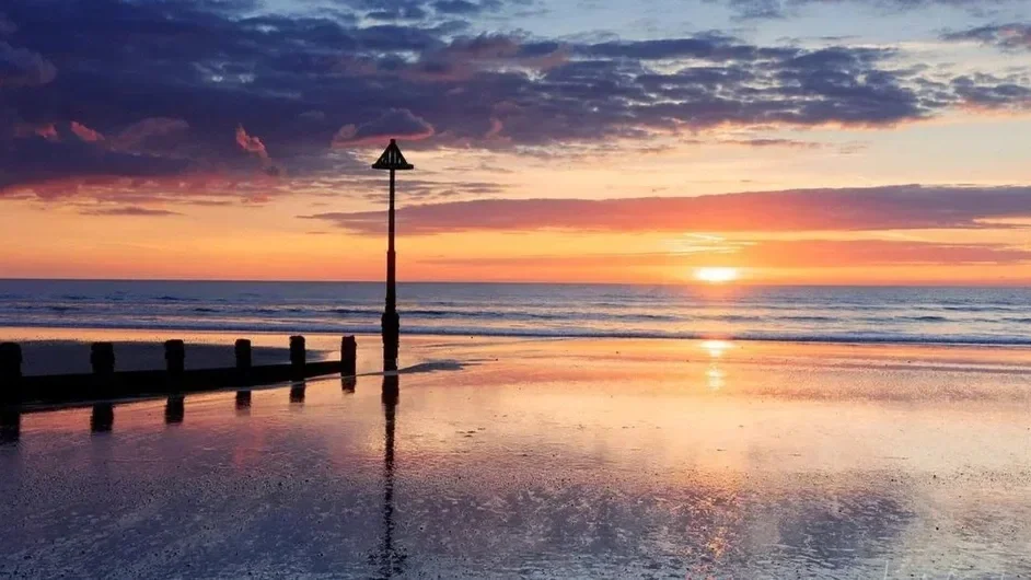 Sunset over a calm beach with reflected sky and clouds, a wooden pier, and a warning or navigation sign in the sand.