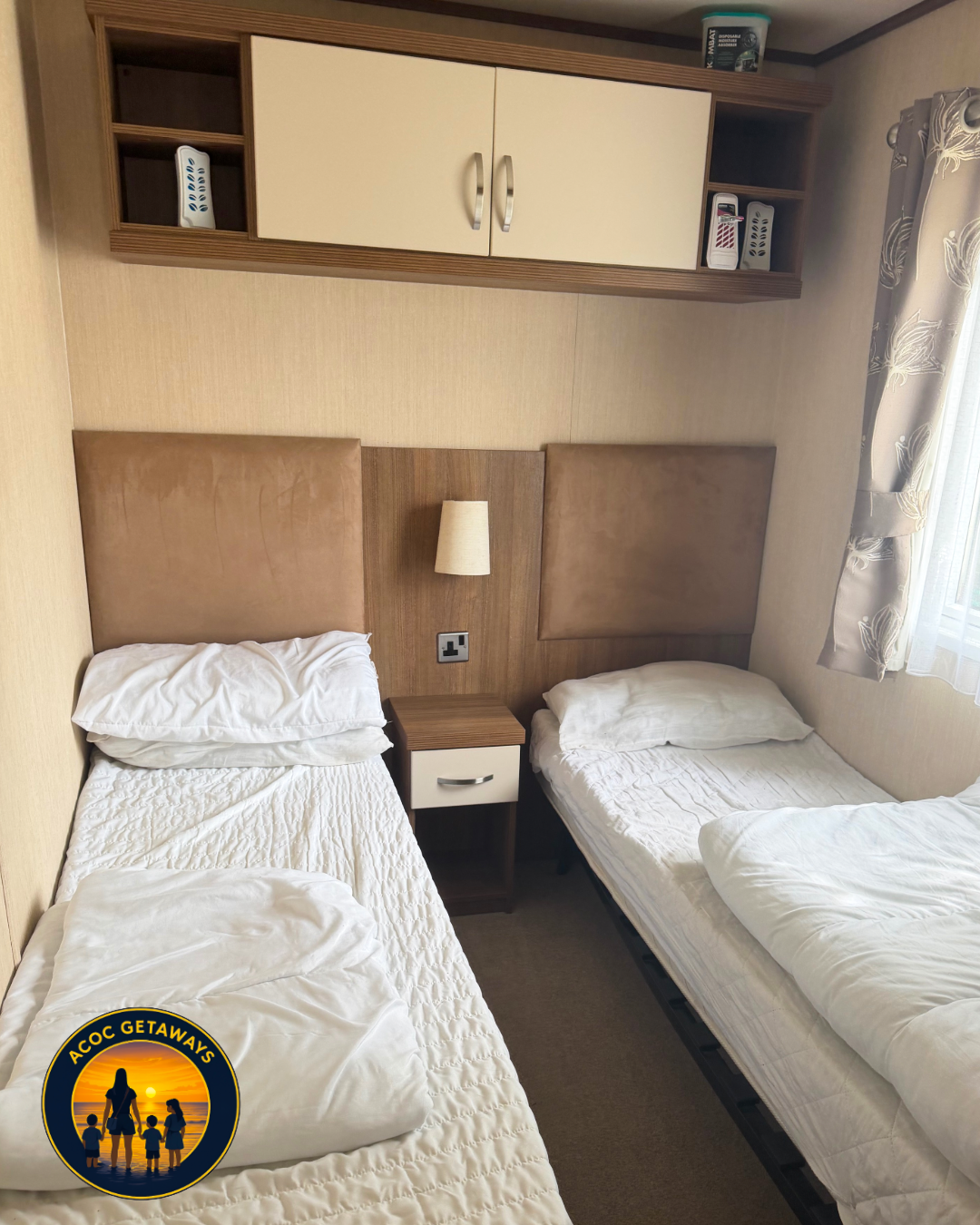 Room with two single beds, a nightstand between them, a wall-mounted lamp, and beige walls with a window covered by curtains featuring a leaf pattern.