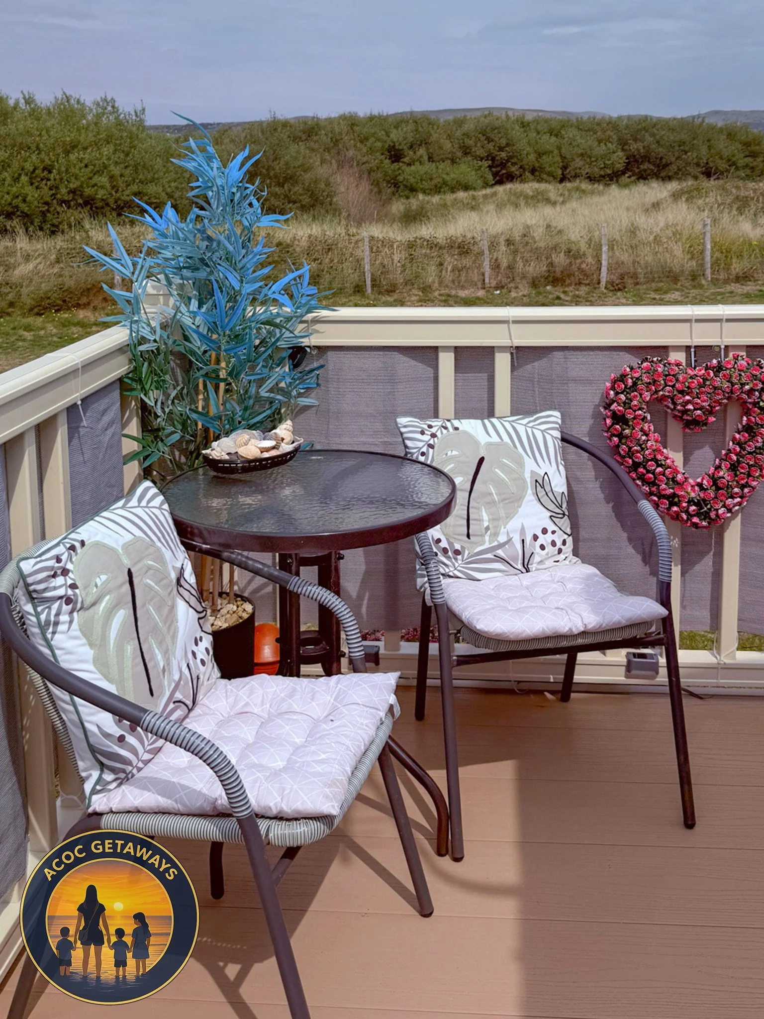 A cozy balcony with two wicker chairs with pillows, a small round glass-top table, decorative plants, a pink heart-shaped flower arrangement on the railing, and a view of a grassy field and distant hills.