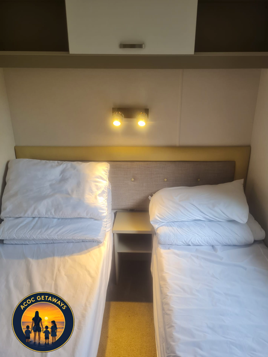 A hotel room featuring two single beds with white bedding and pillows, a beige upholstered headboard, a small white nightstand in between, and a double light fixture above the headboard illuminating the beds.