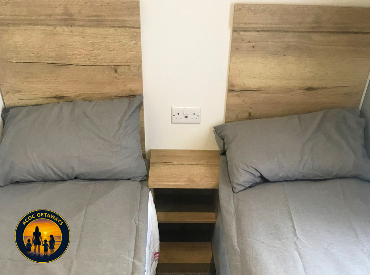 Two beds with gray bedding and pillows, separated by a wooden nightstand, with a power outlet on the wall between them. The headboards are made of horizontal wooden planks. There is a logo in the bottom left corner with a sunset, a woman, and childre