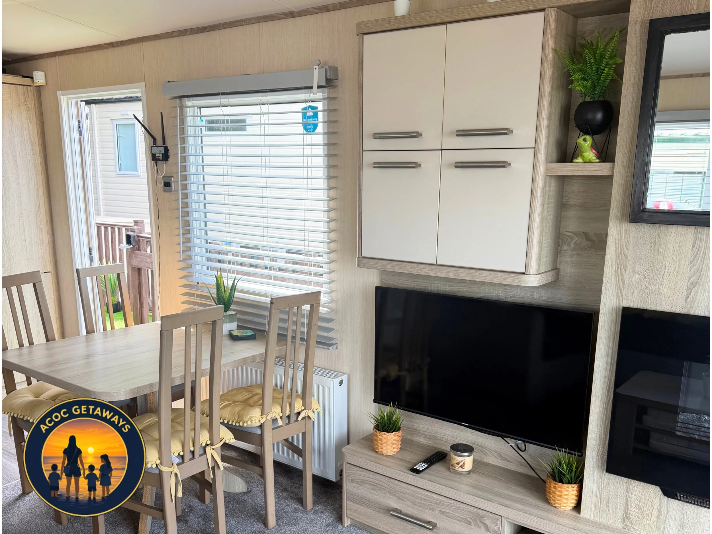 Interior of a living room with beige walls, a dining table with four wooden chairs with yellow cushions, a window with white blinds, a cabinet with closed compartments, a flat-screen TV, small potted plants, and decorative items like a black vase wit
