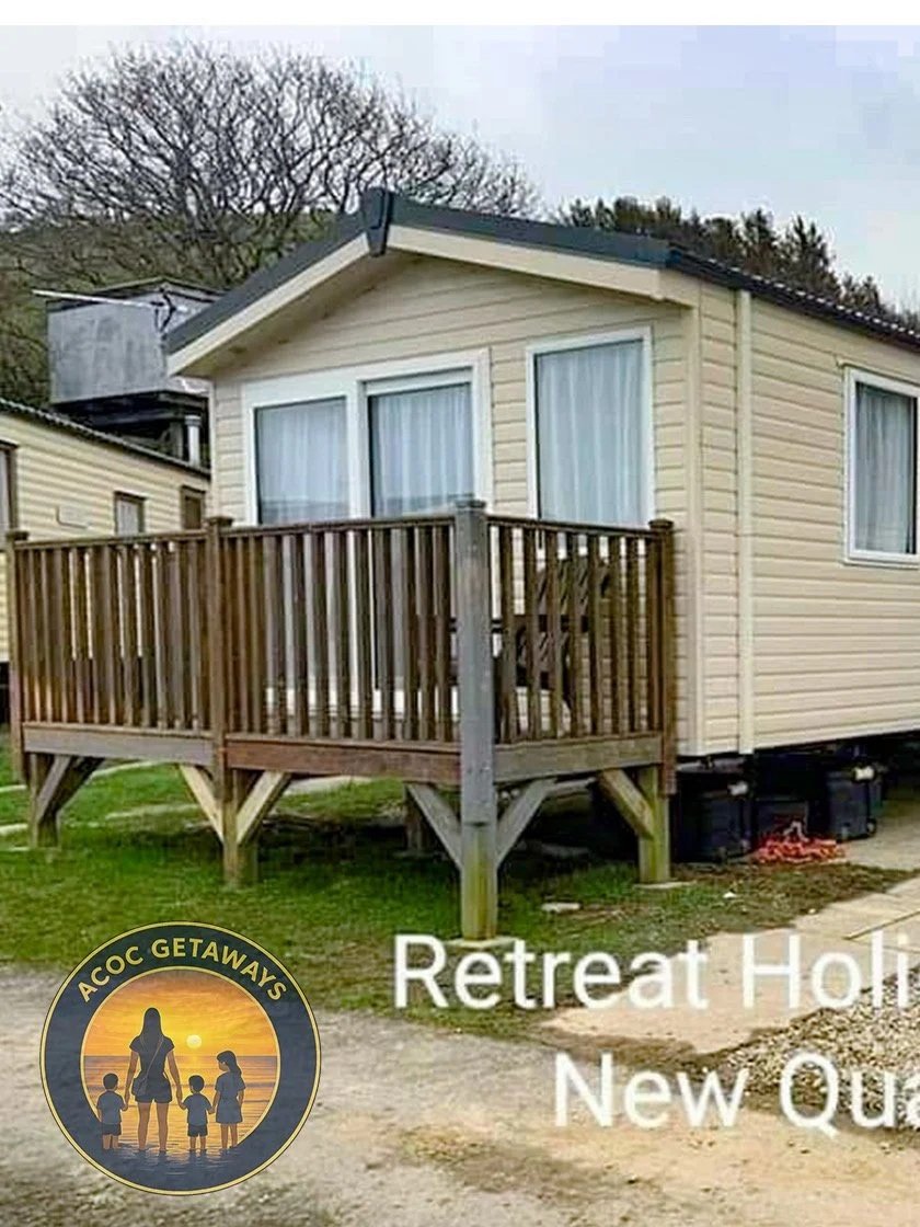 A small beige house with a wooden deck, two large windows with white curtains, and a gabled roof. There are trees in the background and trash bins underneath the deck.