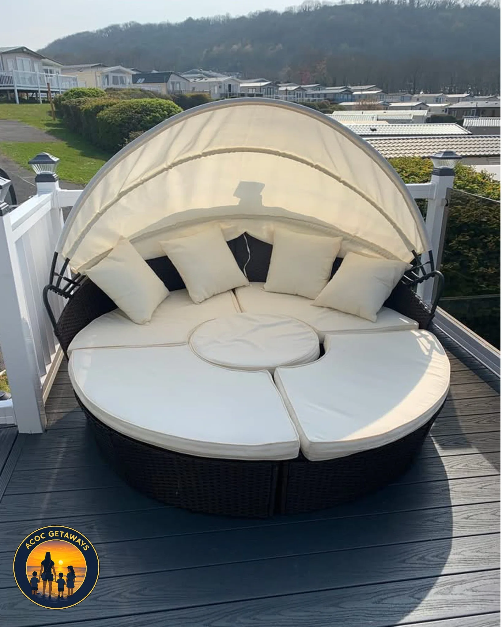 Round outdoor patio furniture with a curved canopy, cream-colored cushions, and pillows, on a wooden deck with a white railing, overlooking houses and hills in the background.