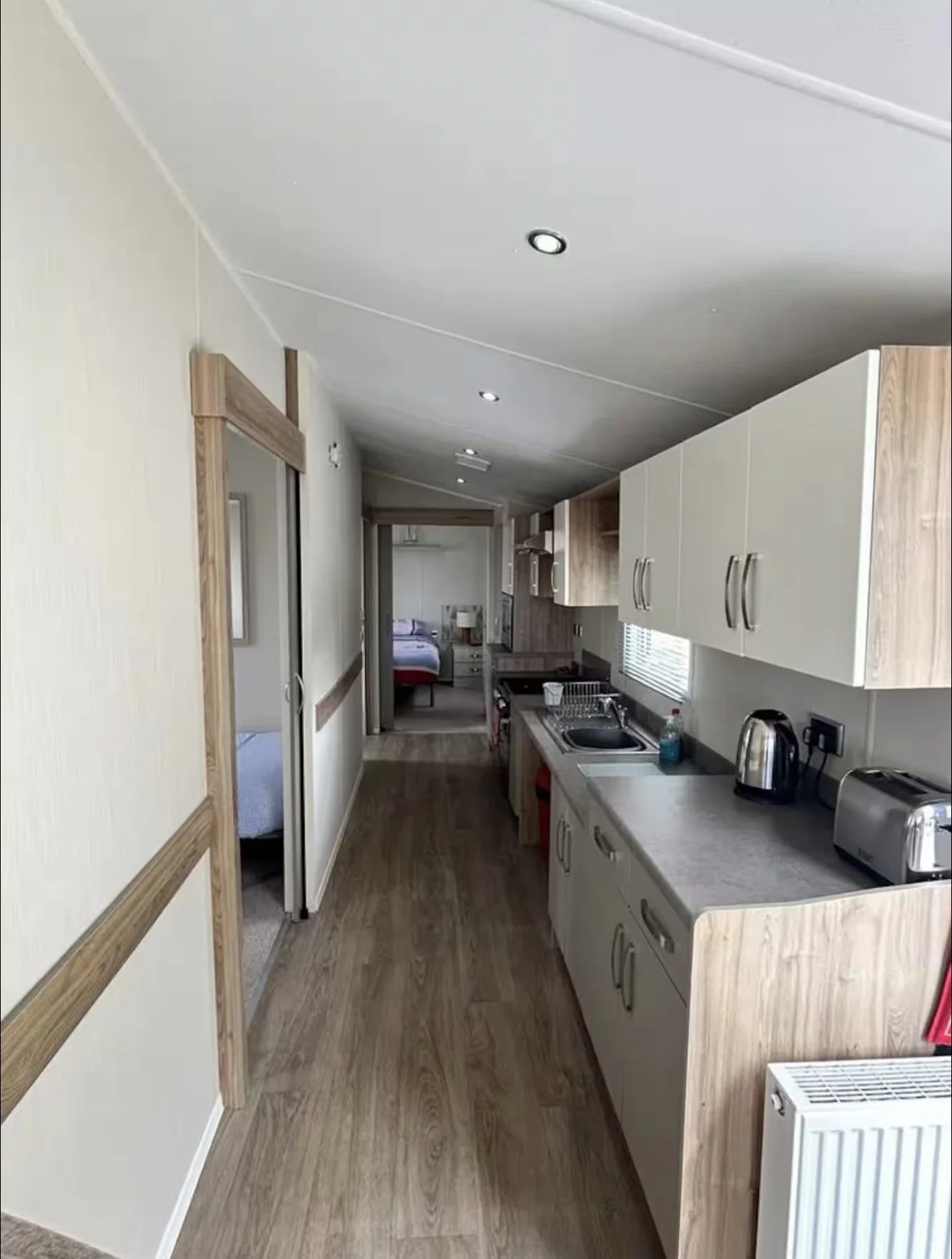 A narrow kitchen with white cabinets, a countertop with a sink, and small appliances, leading to a bedroom with a bed and nightstand in the background.