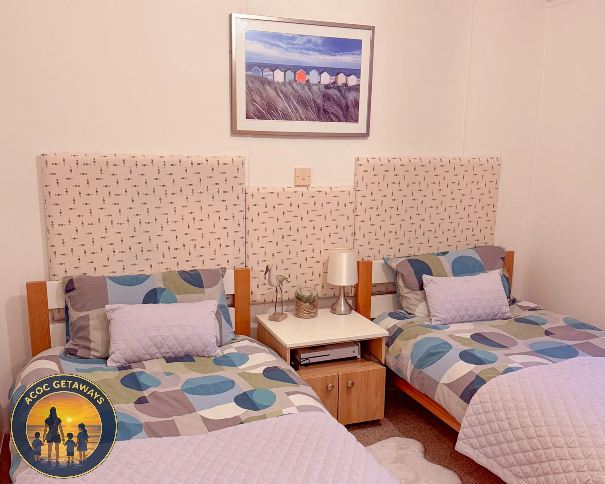 A cozy bedroom with two beds that have patchwork quilt blankets and patterned pillows, separated by a nightstand with a lamp, a small decorative bird, and a basket. There is a framed photo of colorful beach huts above the beds.