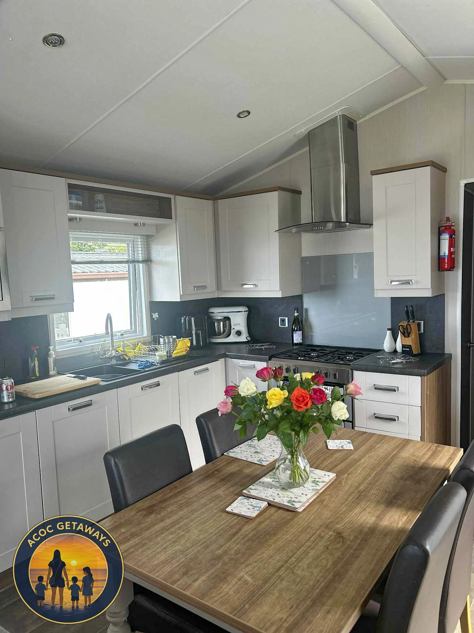 Kitchen with white cabinets, black countertops, a stainless steel range hood, a window above the sink, a dining table with a vase of multicolored roses, and matching napkins.