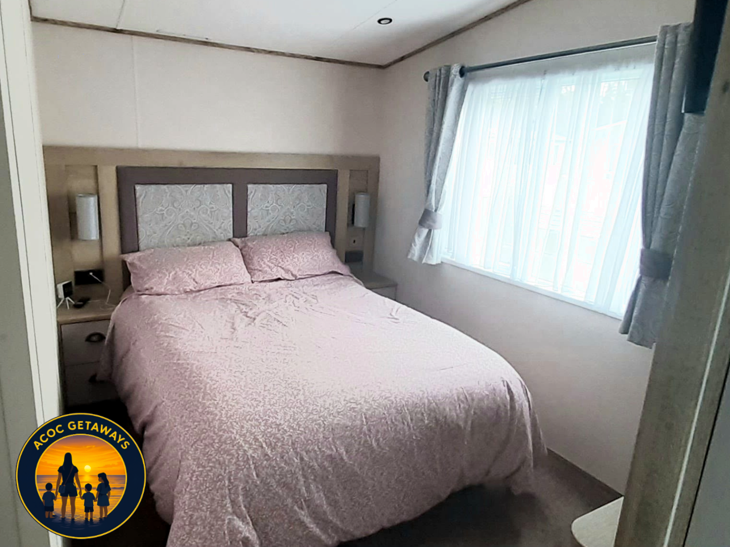 A neatly made bed with pink bedding and matching pillows in a cozy bedroom, featuring a window with curtains and bedside tables with lamps on each side.