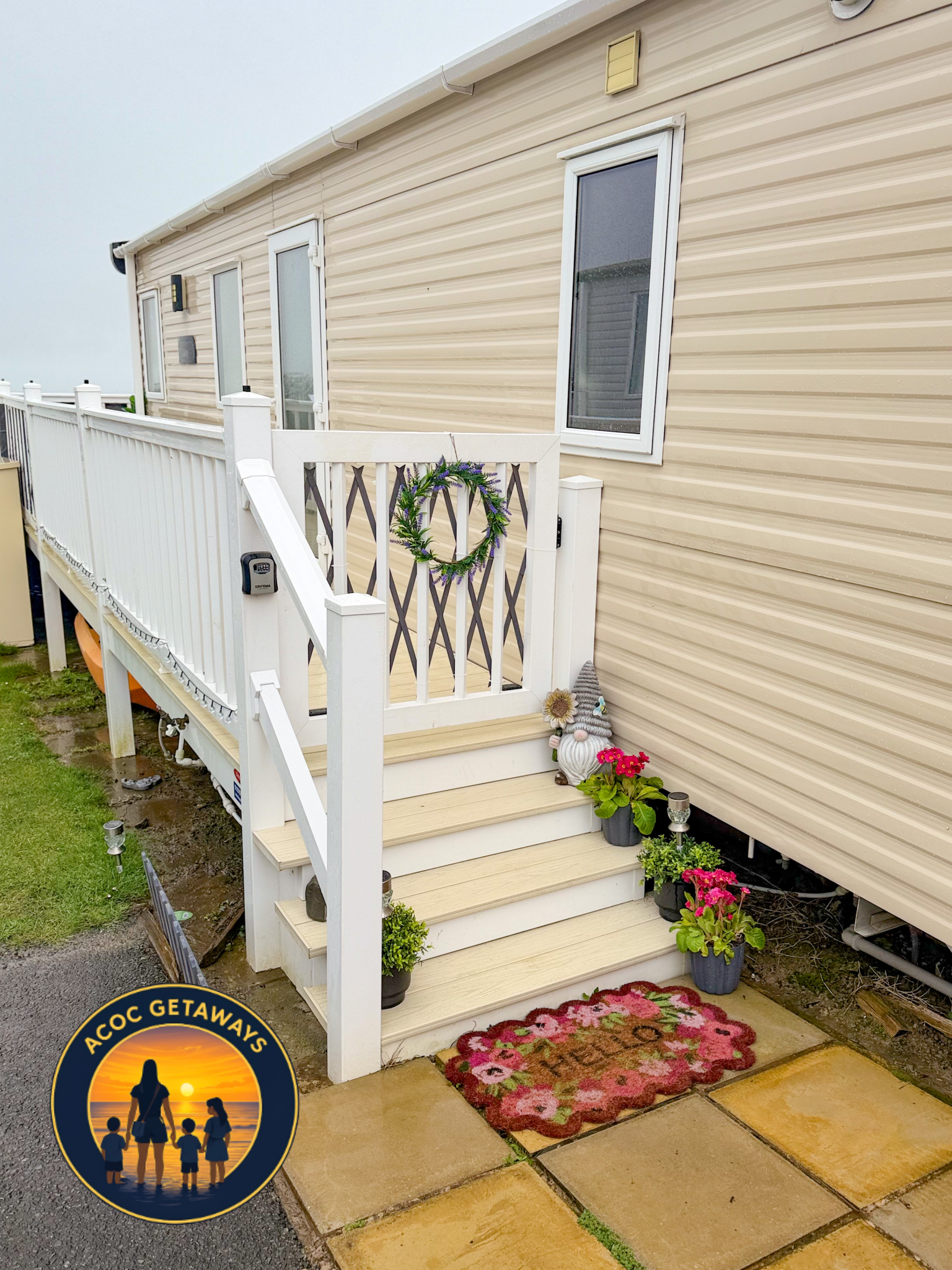 A beige mobile home with a small white porch decorated with a green wreath, potted flowers, and garden gnome figurines, situated on a concrete and stone patio with a colorful floral rug and small solar lights.