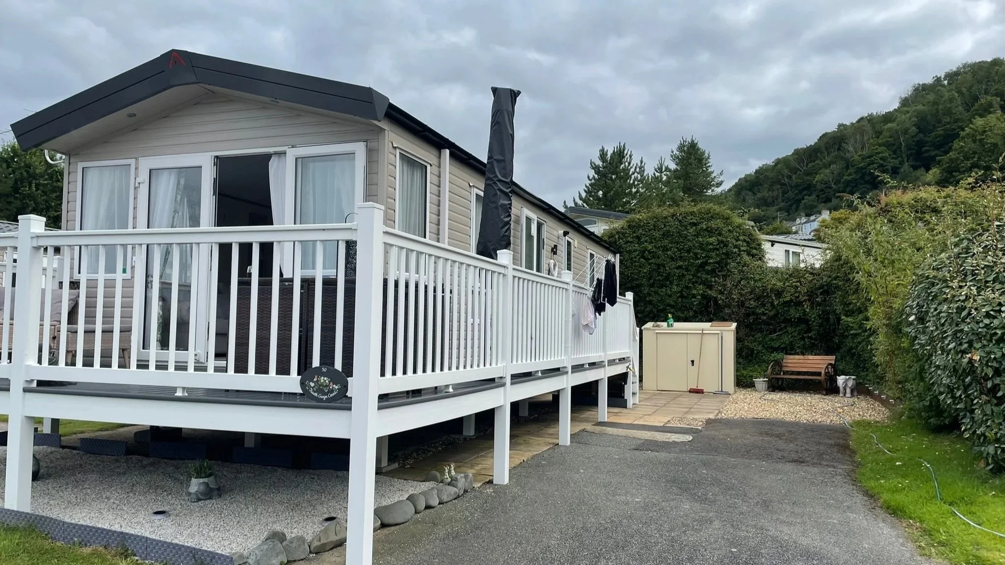 A mobile home with a raised white deck, outdoor clothes hanging to dry, a small garden shed, and a bench with potted plants near a wooded hillside.