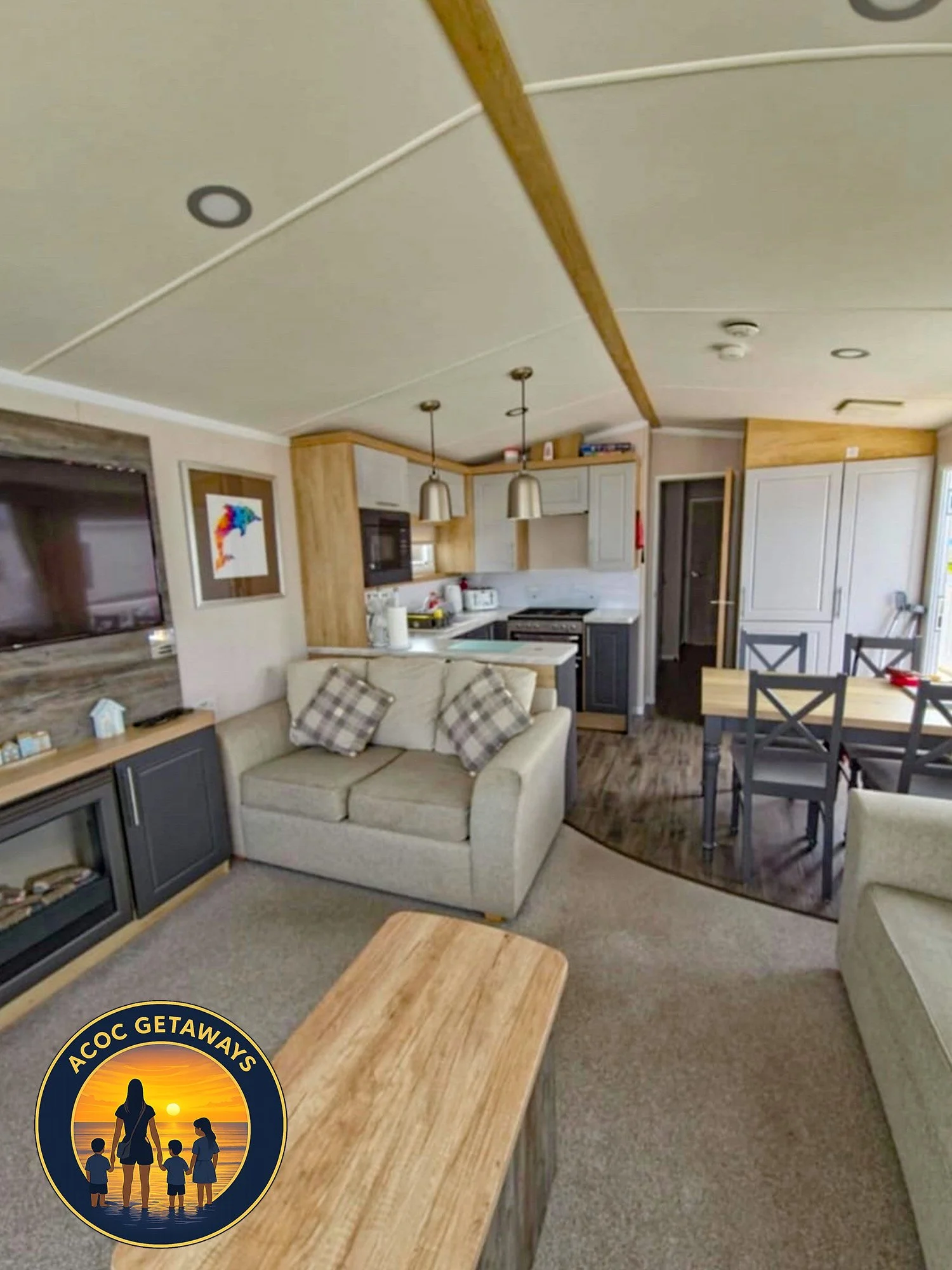 Interior view of a cozy living room and kitchen area in a vacation rental, featuring beige sofas with plaid pillows, a wooden coffee table, a flat-screen TV, a dining table with chairs, and kitchen cabinets with modern appliances.