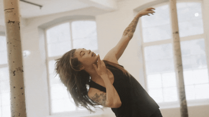 Eine junge Frau tanzt in einem hellen Raum mit großen Fenstern, trägt ein schwarzes Top und hat Tattoos an ihrem Arm.