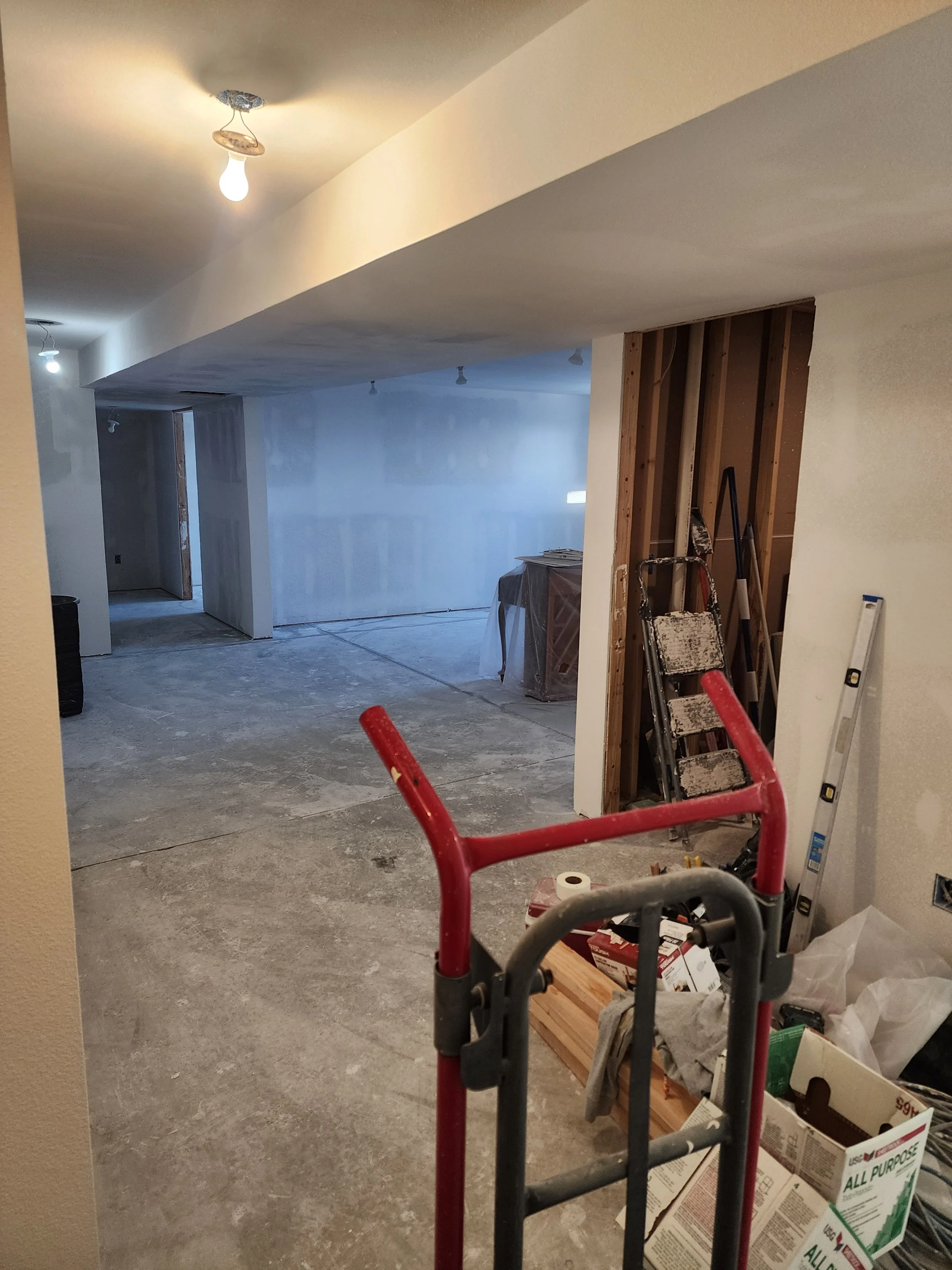 Interior of a house under construction or renovation, with unfinished flooring, construction tools, ladder, and partially installed drywall.