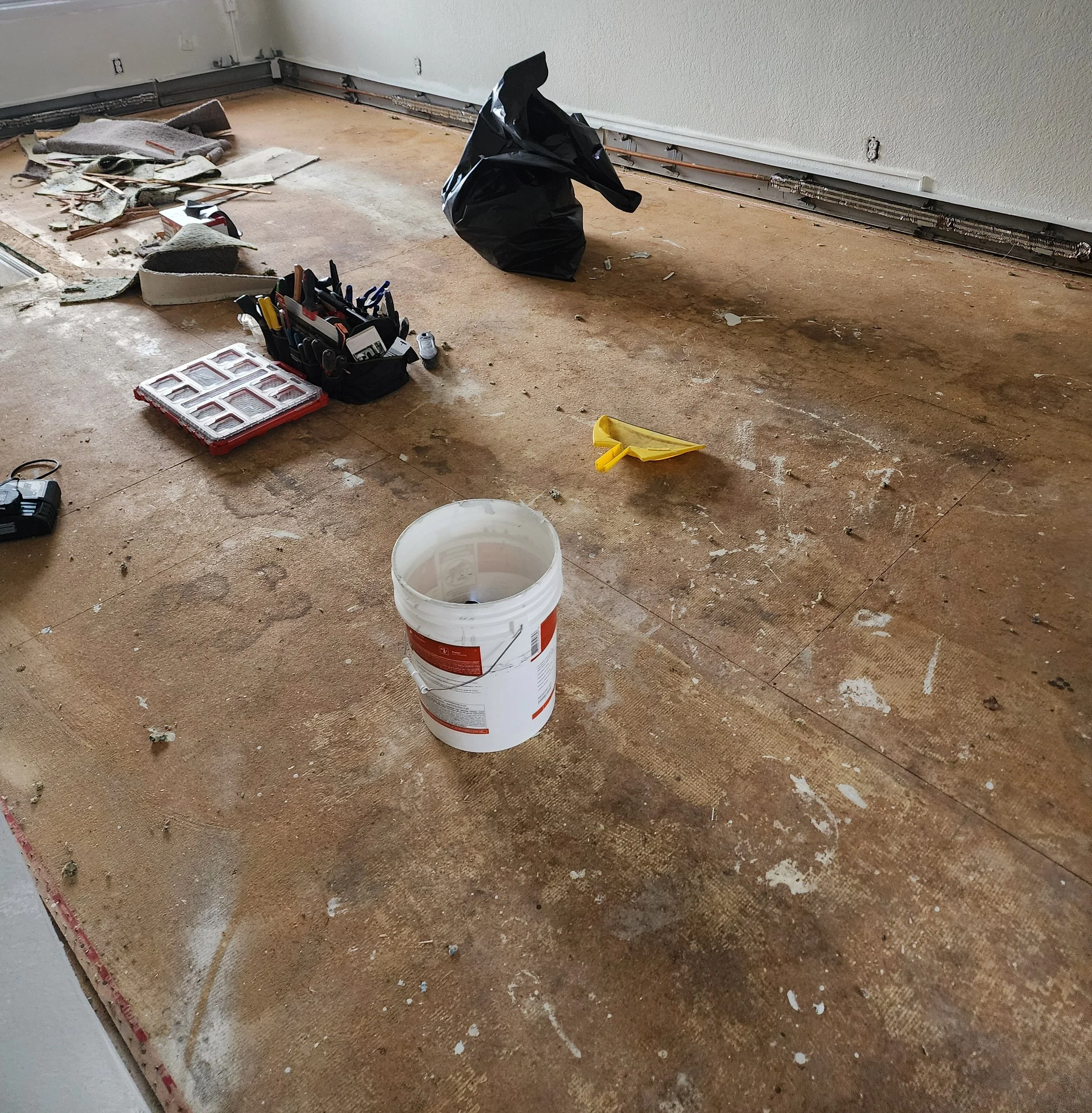A partially renovated room with a large wooden floor, tools, and materials scattered. Includes a white bucket, a yellow dustpan, a black tool bag, a small tool case, and debris from flooring or wall work.